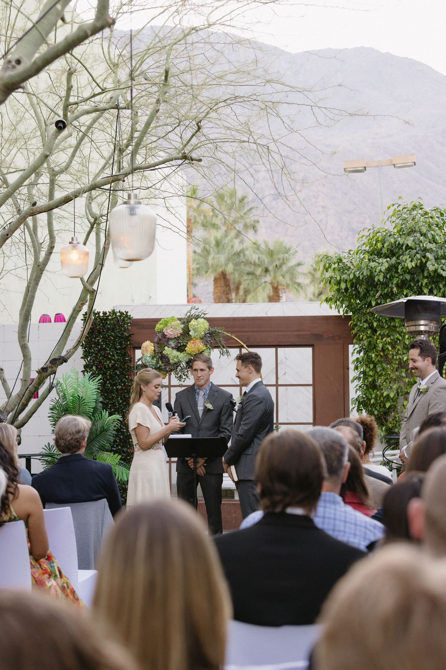 Outdoor wedding ceremony in Palm Springs with couple exchanging vows, mountain backdrop, lush greenery, and seated guests.