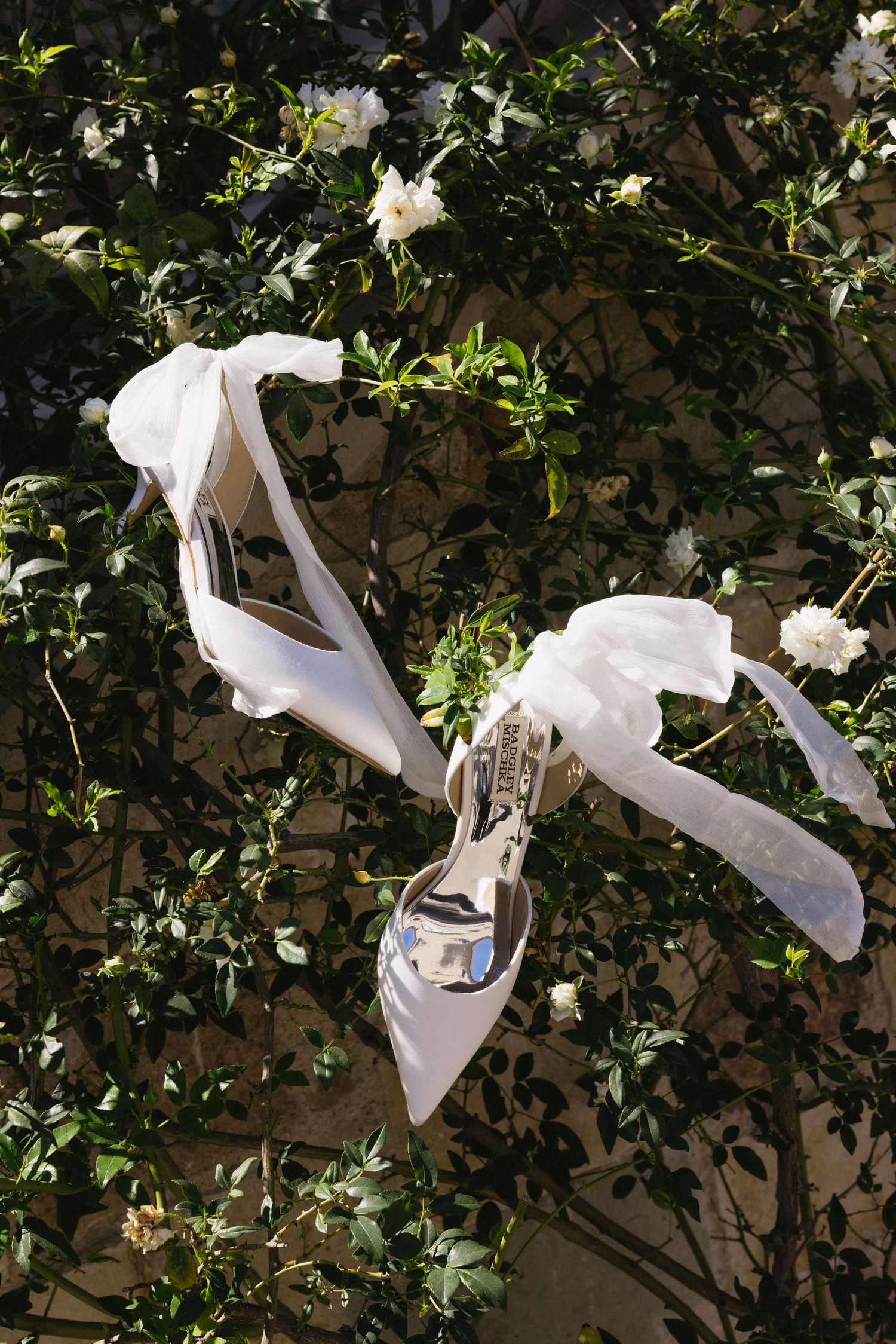 White bridal heels with ribbon bows displayed against lush green foliage and white flowers.