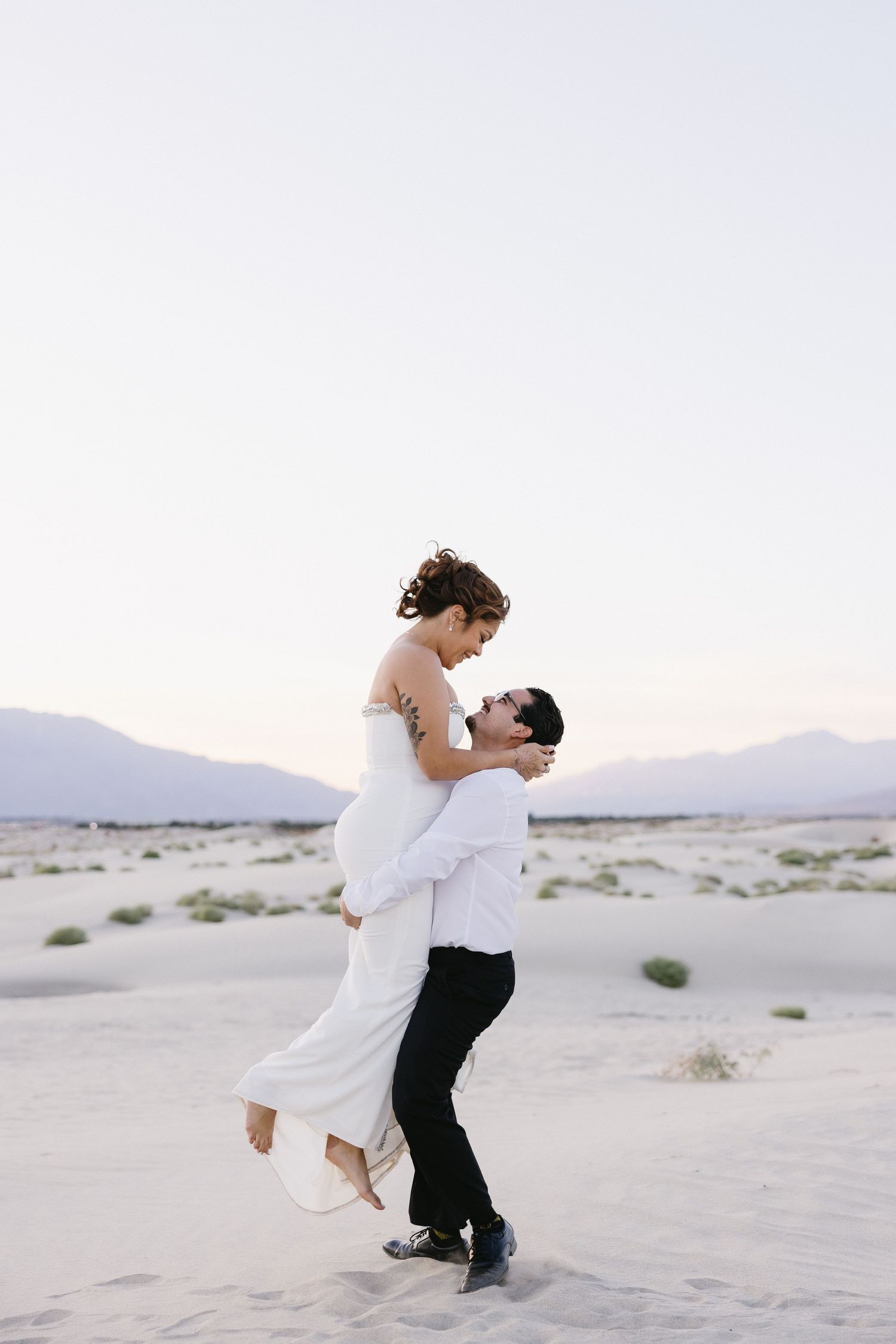 Romantic moment captured as couple embraces on sandy dunes during desert photoshoot.