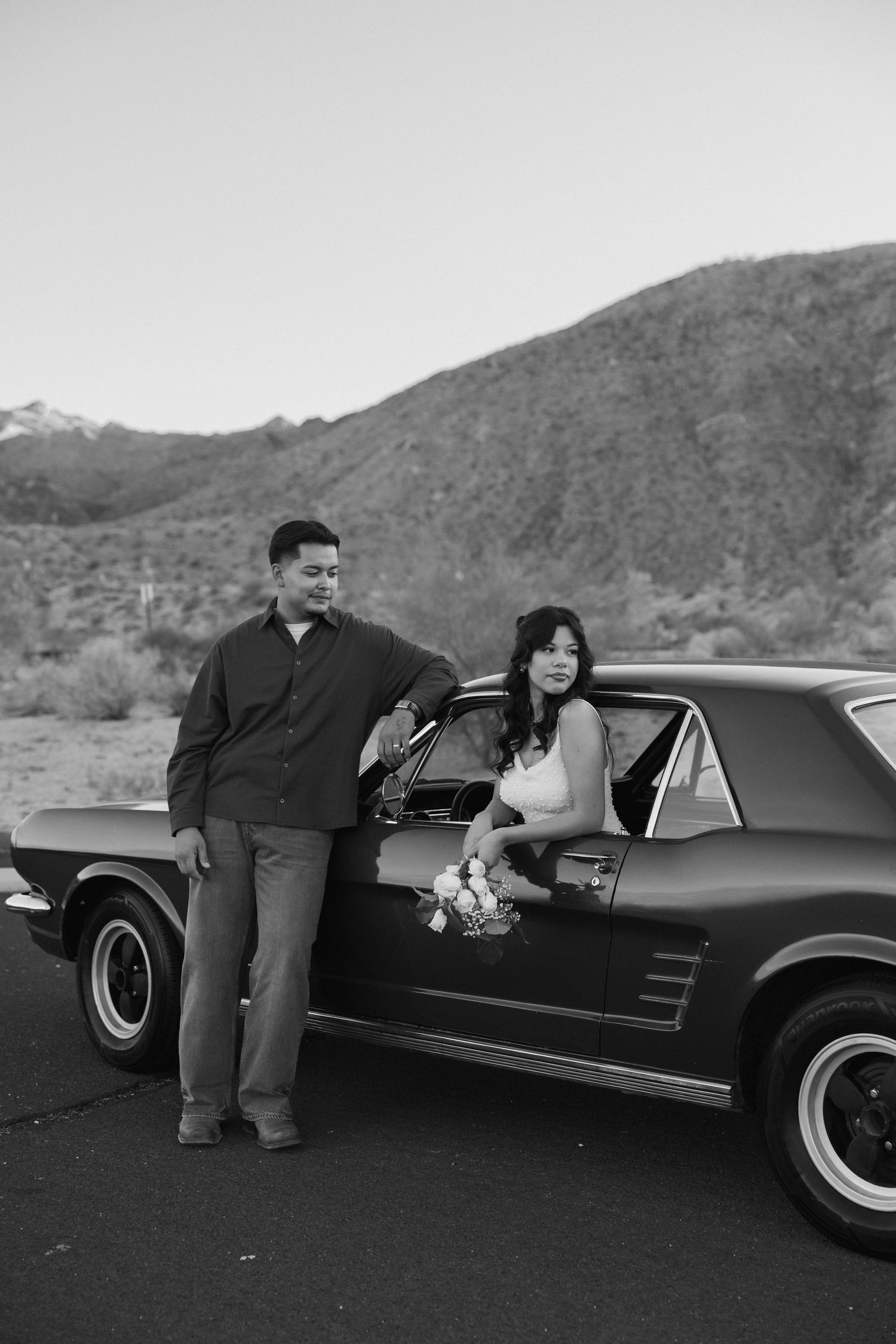 Couple sharing intimate moment by classic black car with desert mountains behind them.