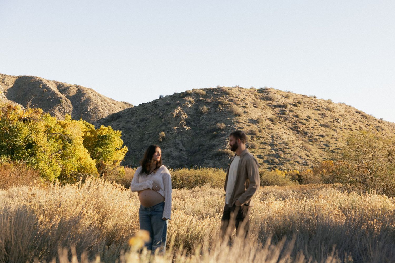 Two people stand together in a desert field with mountains and golden trees in the background.