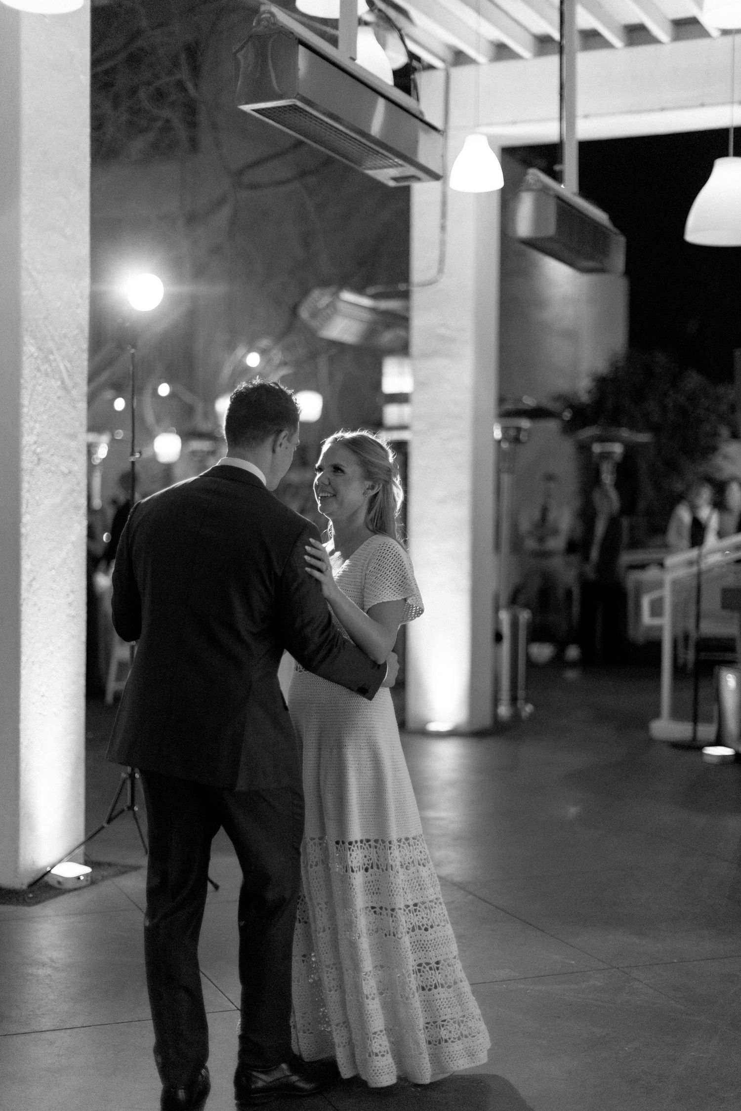 Black and white photo of a couple sharing their first dance at a wedding reception under warm evening lights.