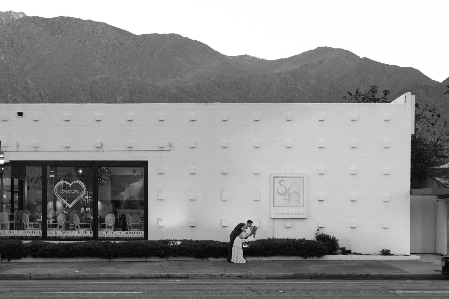 Black and white photo of a couple kissing outside a white building with mountains in the background.