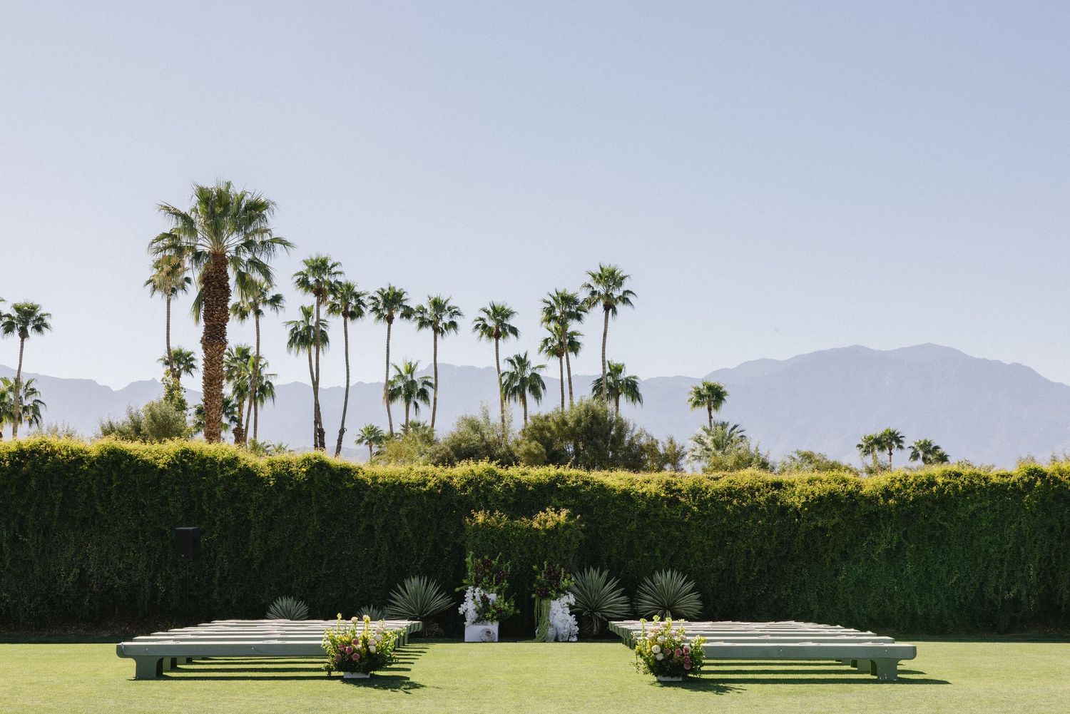 Outdoor wedding ceremony setup with green benches and floral arrangements on manicured lawn with palm trees and mountains.