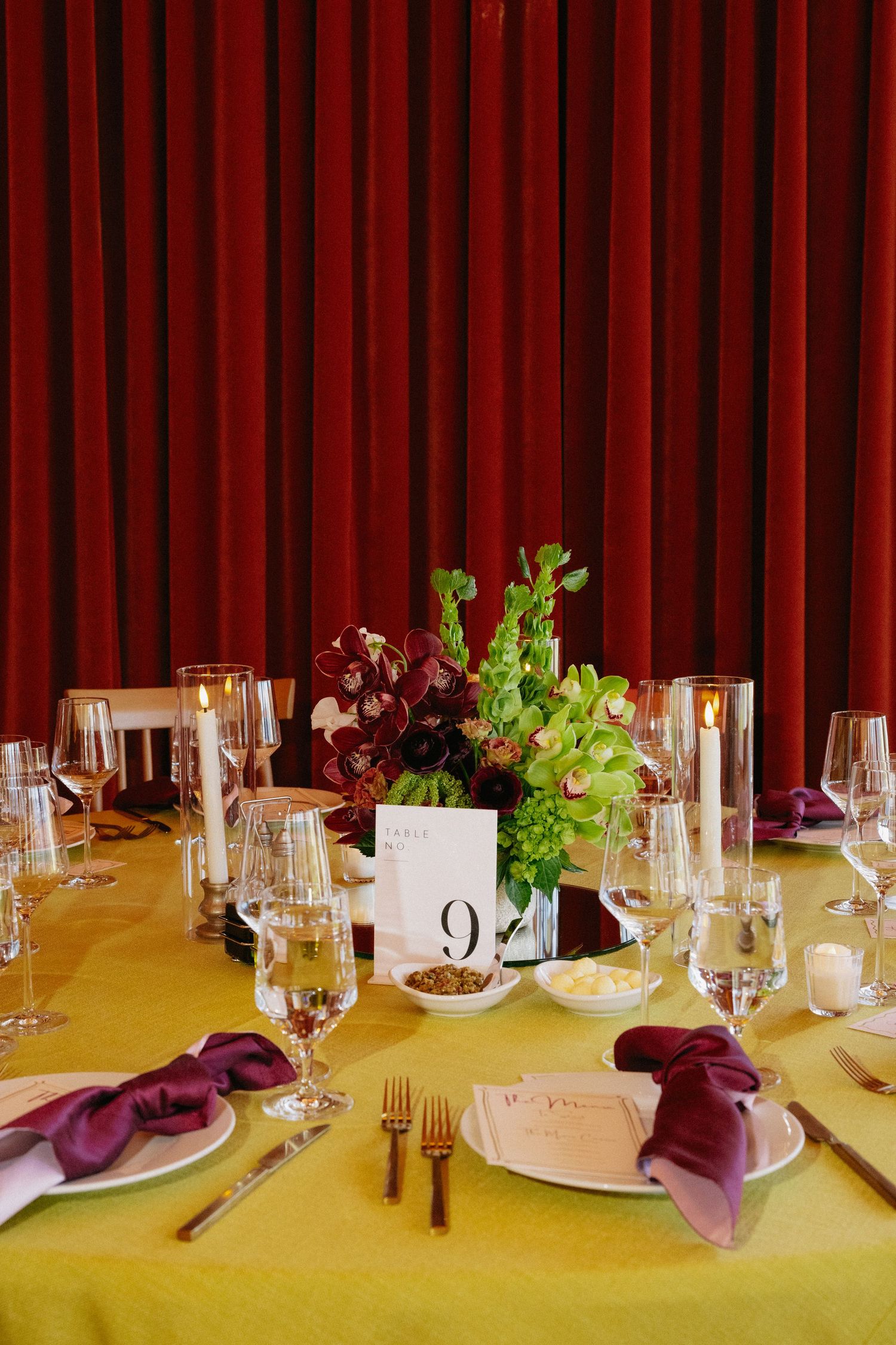Elegant wedding reception table with yellow linen, purple napkins, gold cutlery, floral centerpiece, and red velvet backdrop.