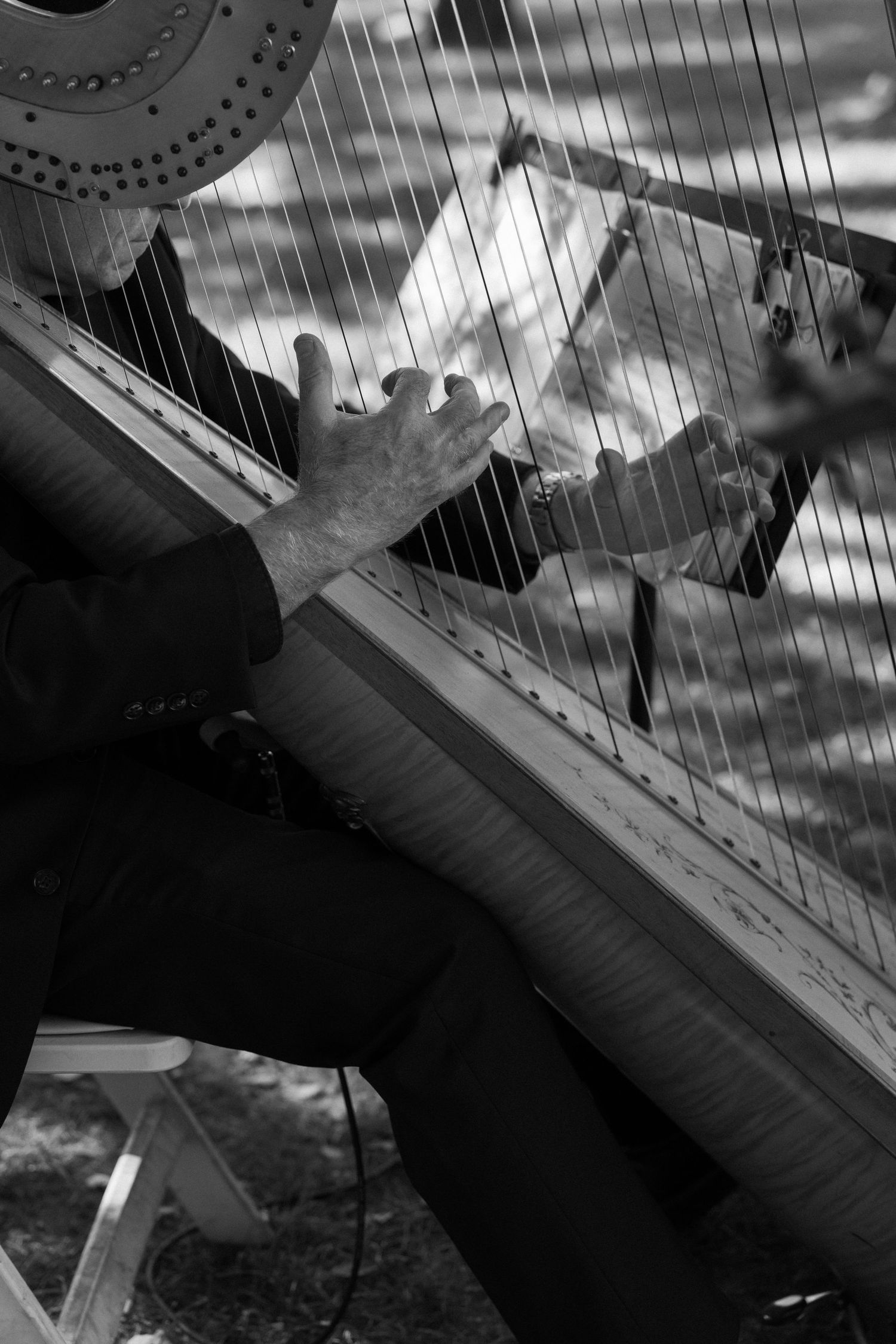 Black and white photo of a musician playing a harp outdoors, fingers plucking strings with sheet music visible.