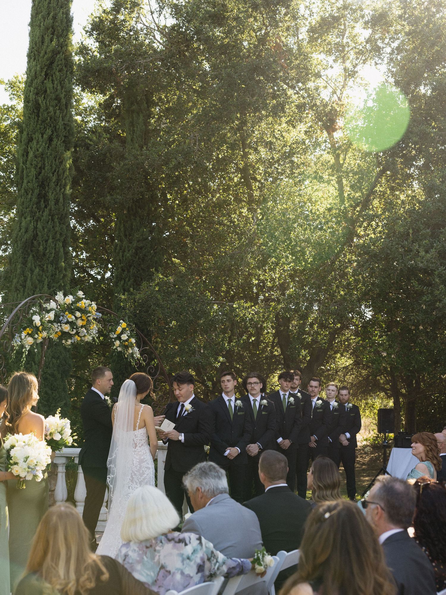 Outdoor wedding ceremony with bride and groom at altar, groomsmen in black suits, lush green trees, and floral arrangements.