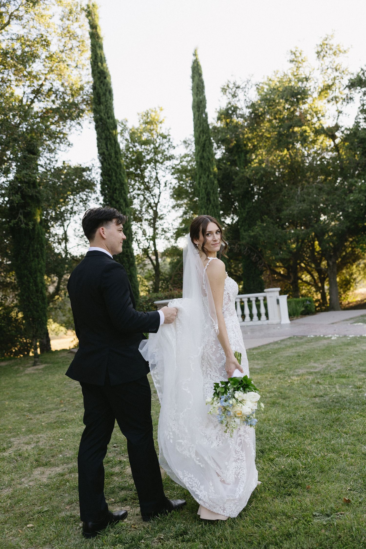 Bride and groom walking outdoors on green lawn, groom holding wedding dress train, bride carrying white bouquet.