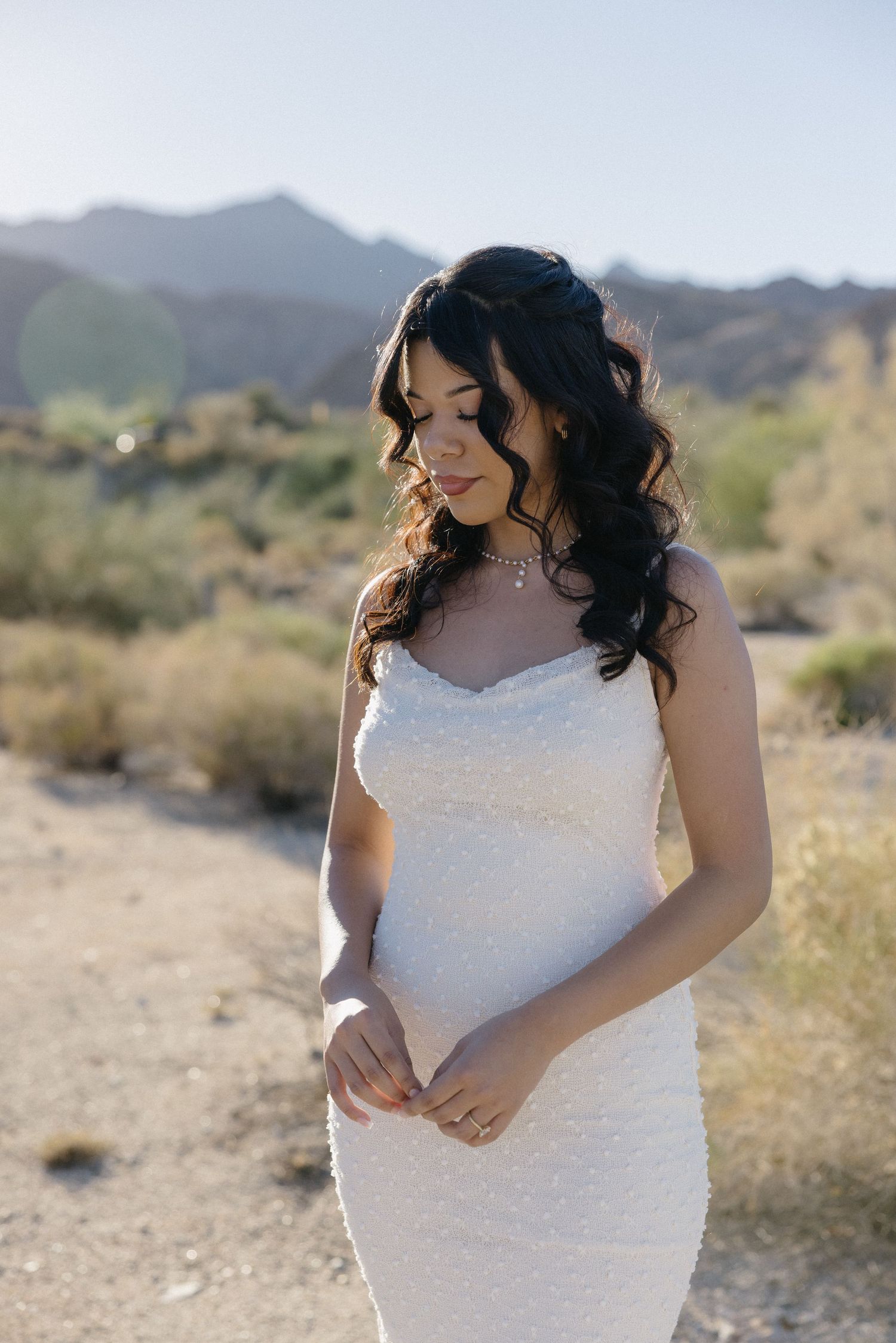 Bride in white lace dress poses in desert landscape with mountains.