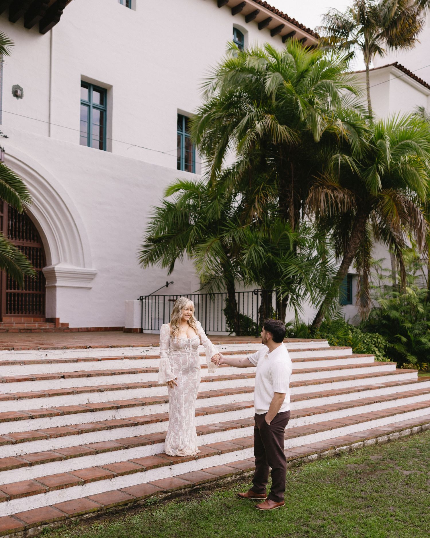 A couple stands on wide stone steps leading up to a white Spanish colonial building with palm trees.