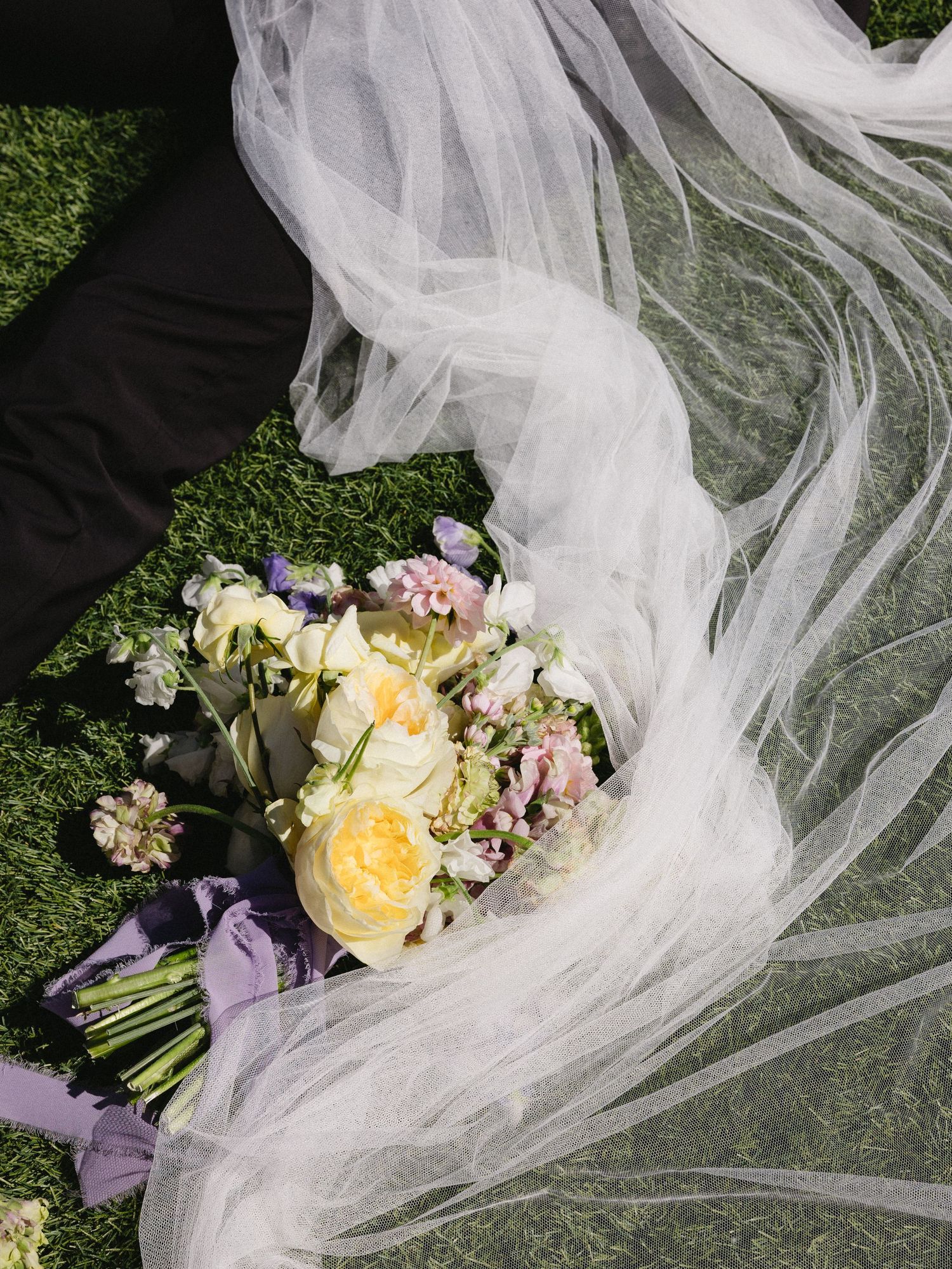 A bridal bouquet of yellow and pink flowers rests on green grass beside a flowing white wedding veil.