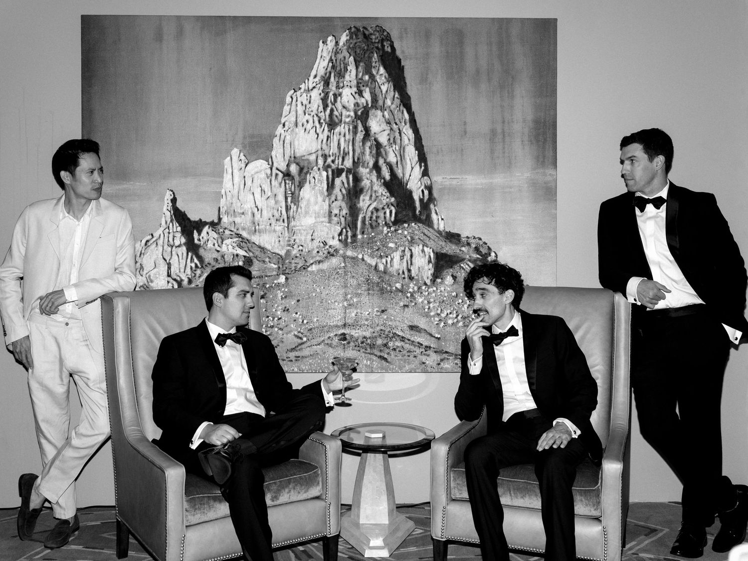 Groomsmen in black tuxedos relaxing in modern lounge with dramatic mountain artwork in black and white photo.