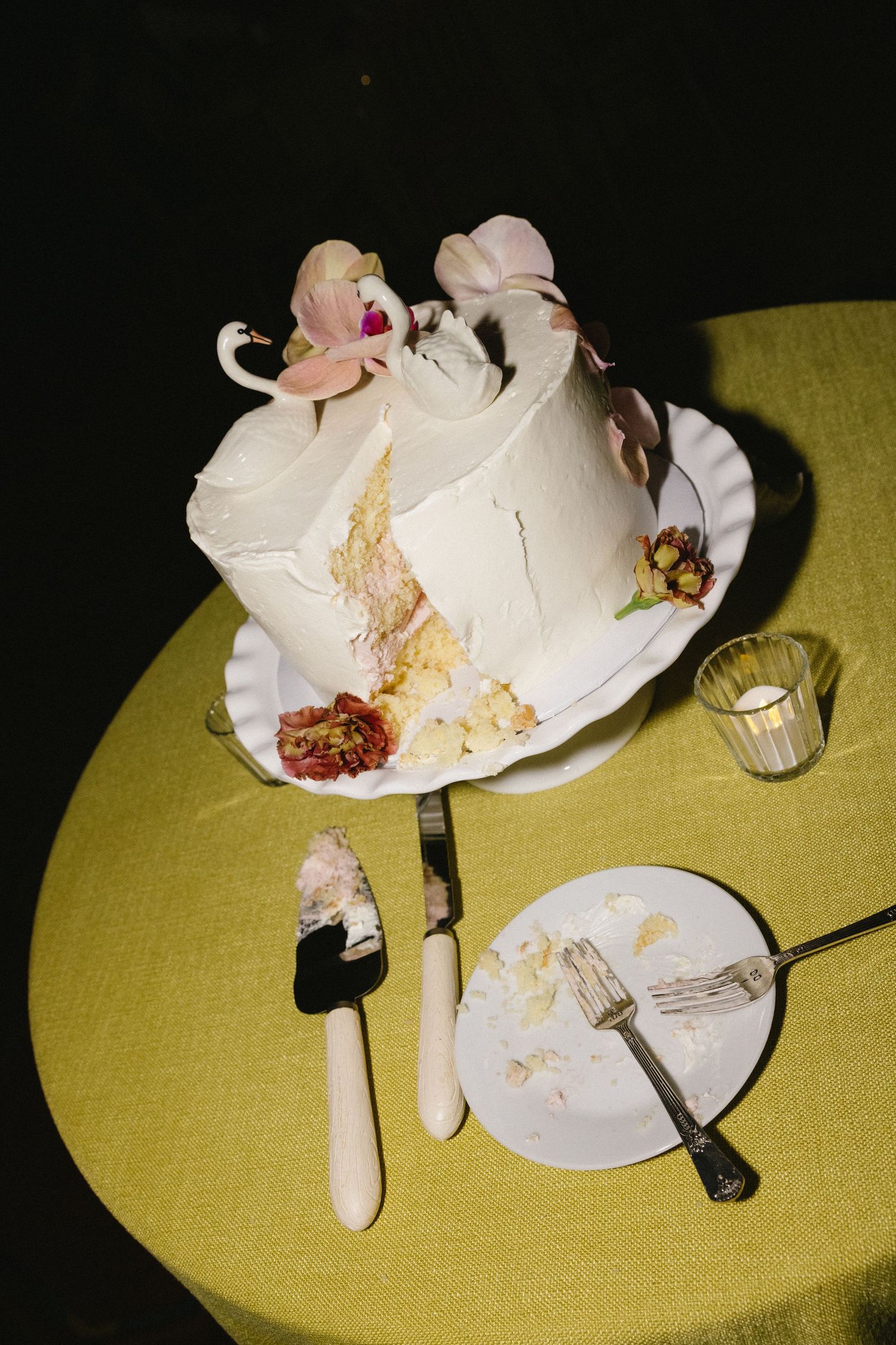 Partially sliced white wedding cake with floral decorations on yellow tablecloth with cake server and plate.