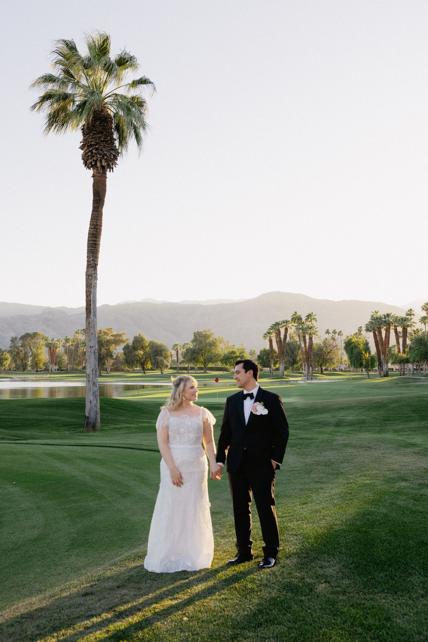 Bride and groom holding hands on lush golf course with palm tree and mountain backdrop on wedding day.