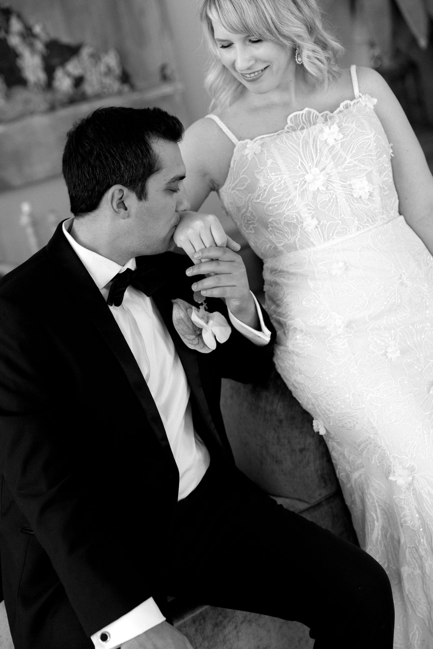 Groom in black tuxedo kisses bride's hand while she smiles in lace wedding dress, black and white photo.