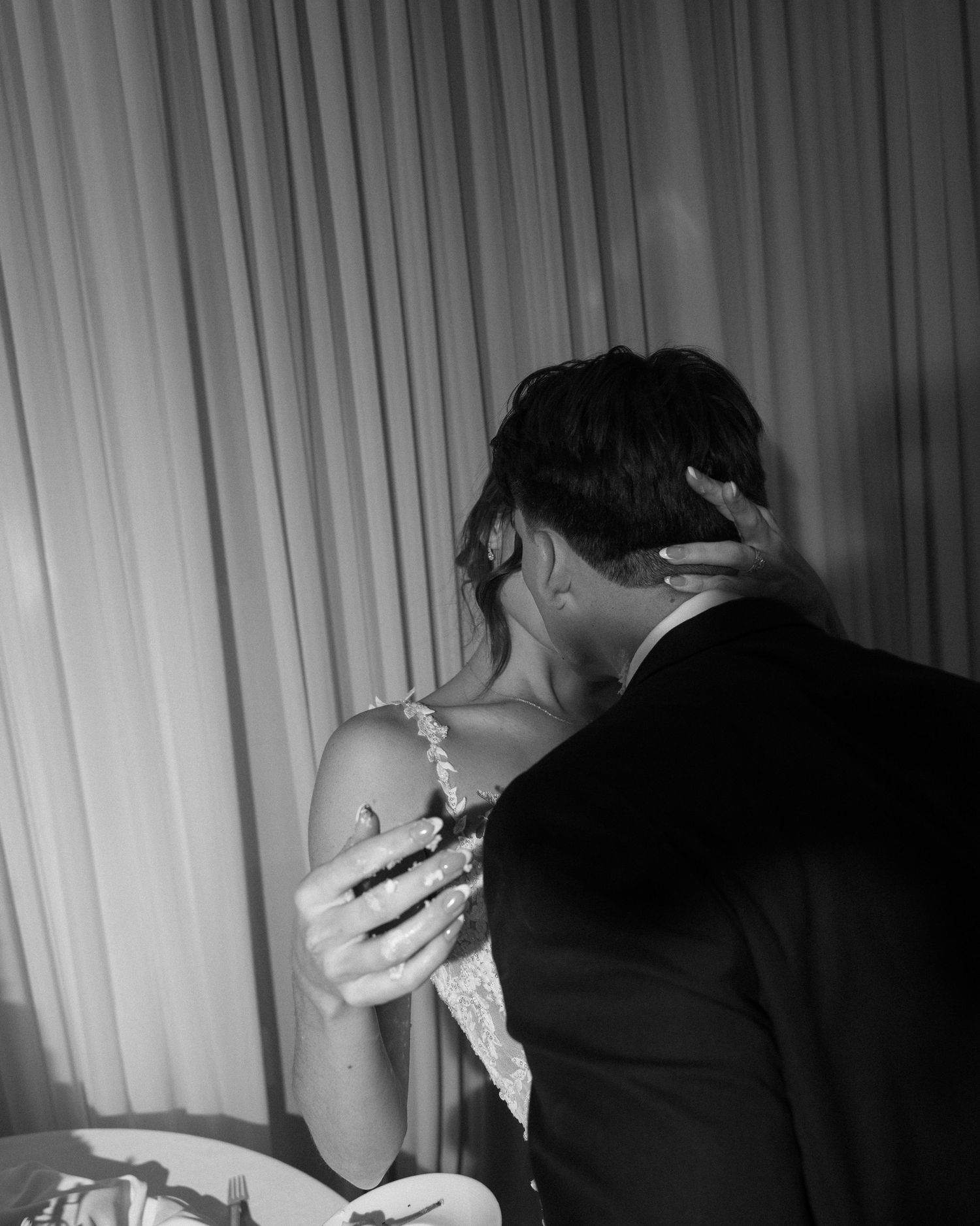 Black and white photo of a couple embracing and kissing at a formal event, bride in lace dress, groom in suit.