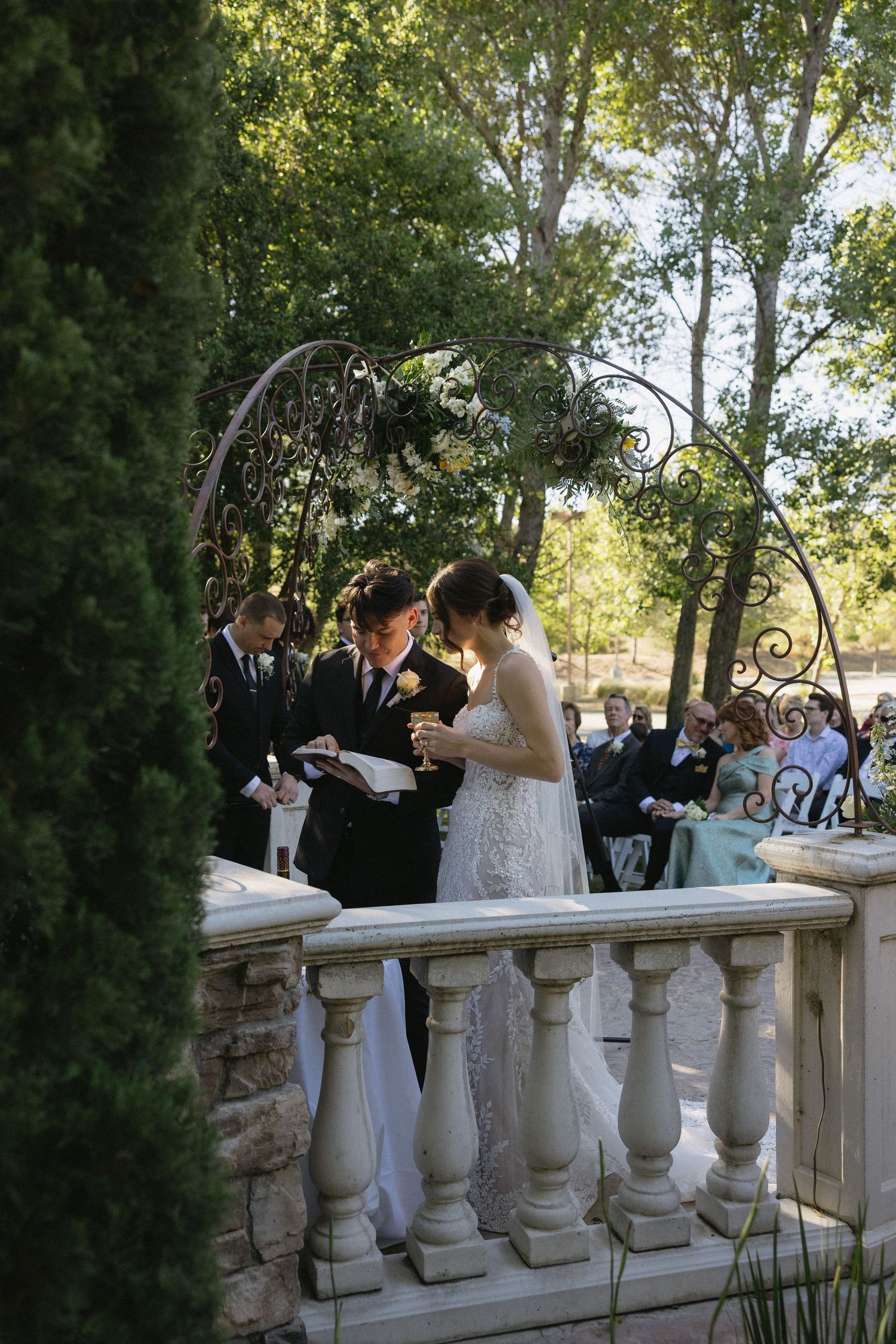 Bride and groom exchange vows at elegant outdoor wedding ceremony on stone balcony with ornate iron arch and lush greenery.