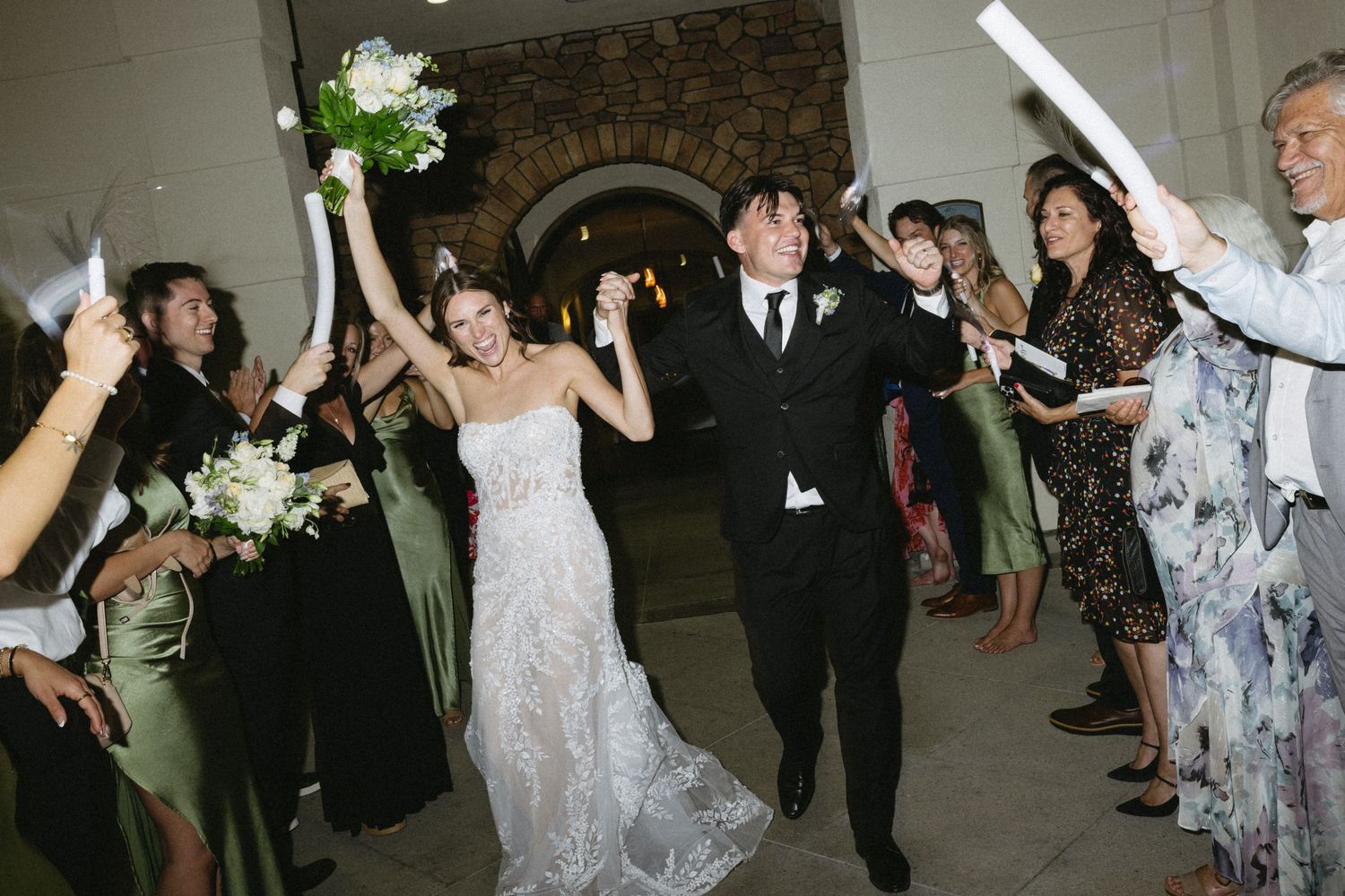 Bride and groom exit wedding reception through cheering guests waving white sparklers and bouquet raised in celebration.