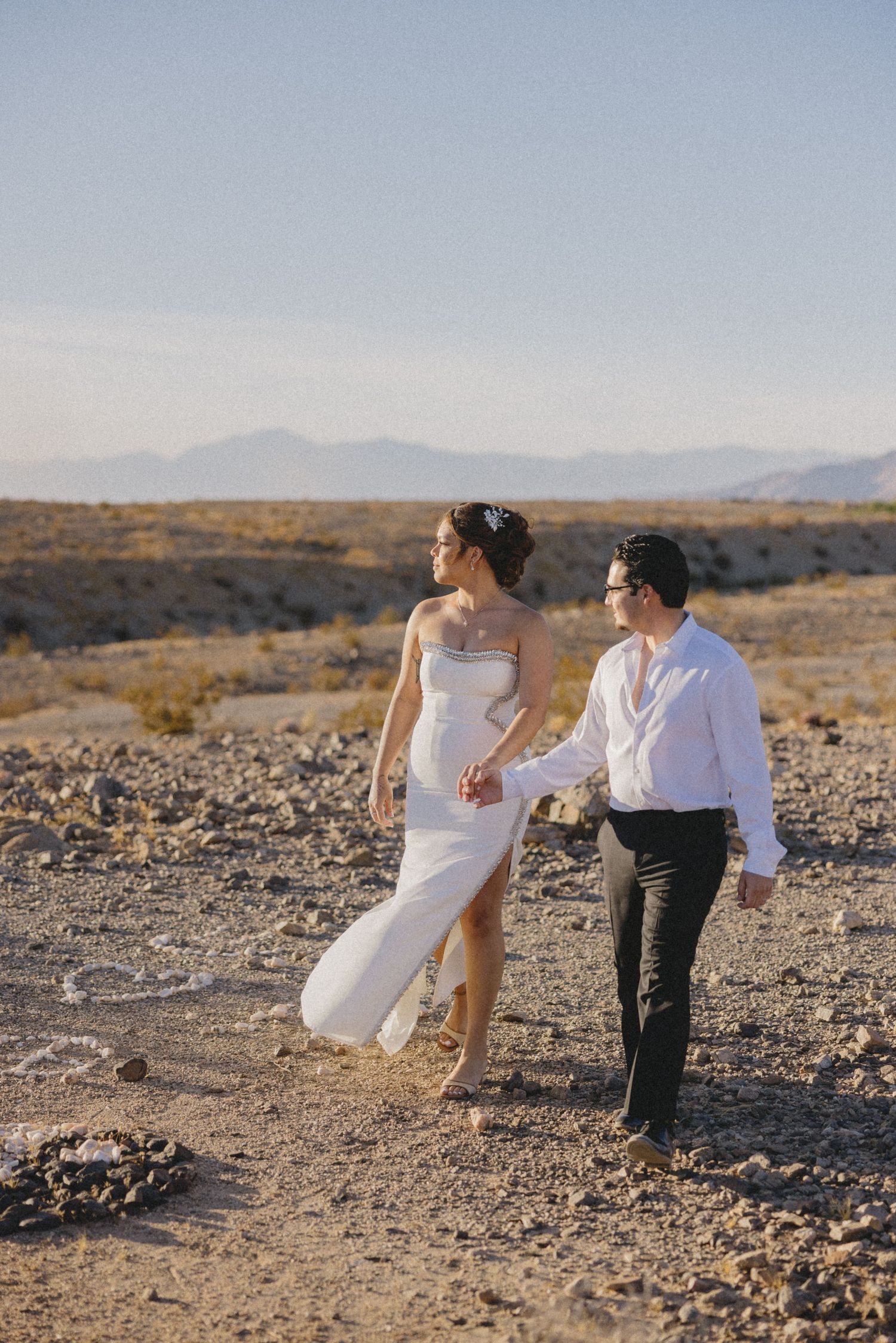 A couple in wedding attire walks together across a desert landscape with mountains in the background at sunset.