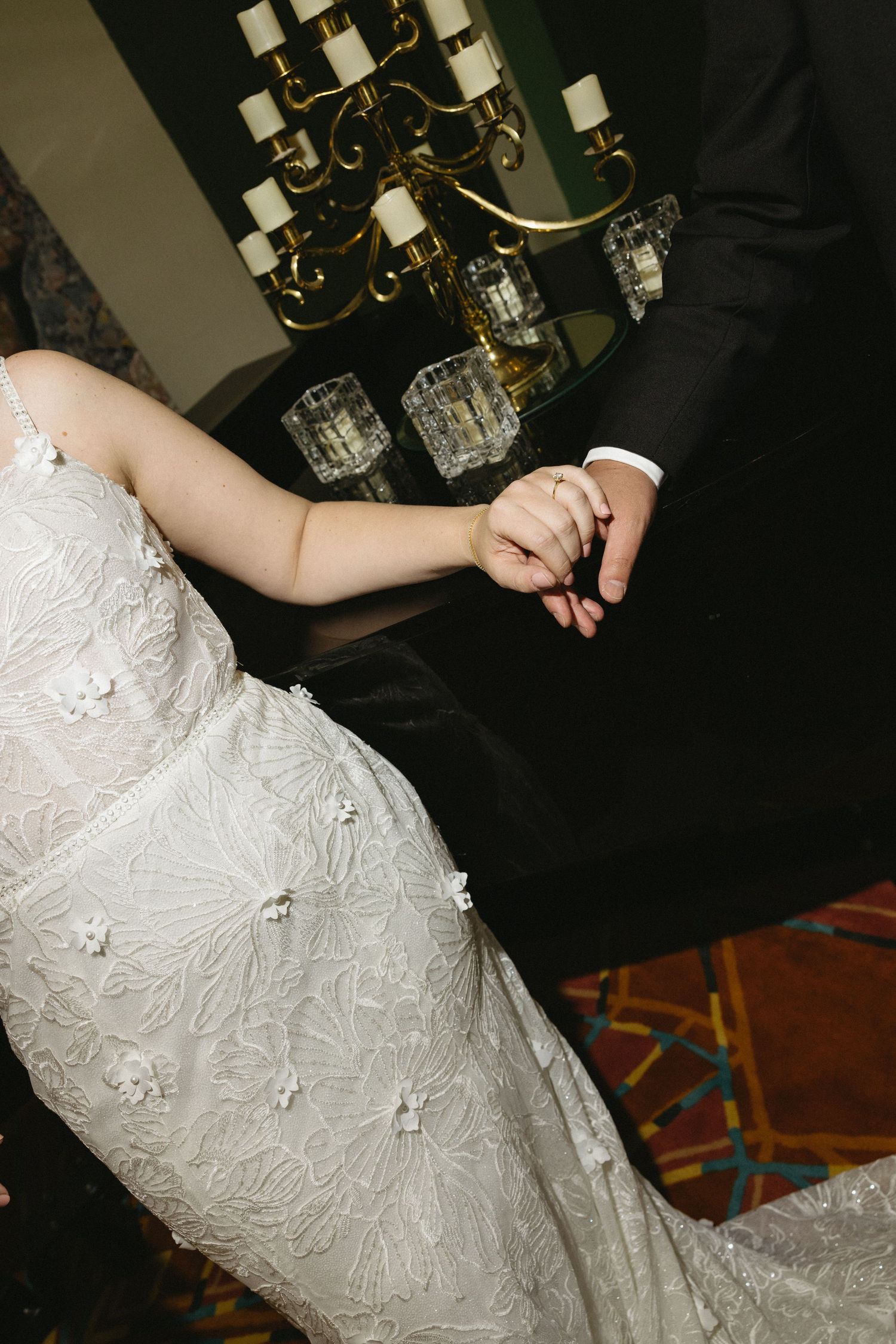 Bride in embroidered lace wedding gown holding hands with groom in black suit near candlelit chandelier.