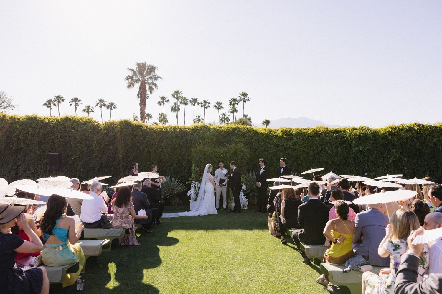 Outdoor wedding ceremony on lush green lawn with guests holding parasols, palm trees, and mountain backdrop in sunny Palm Springs.