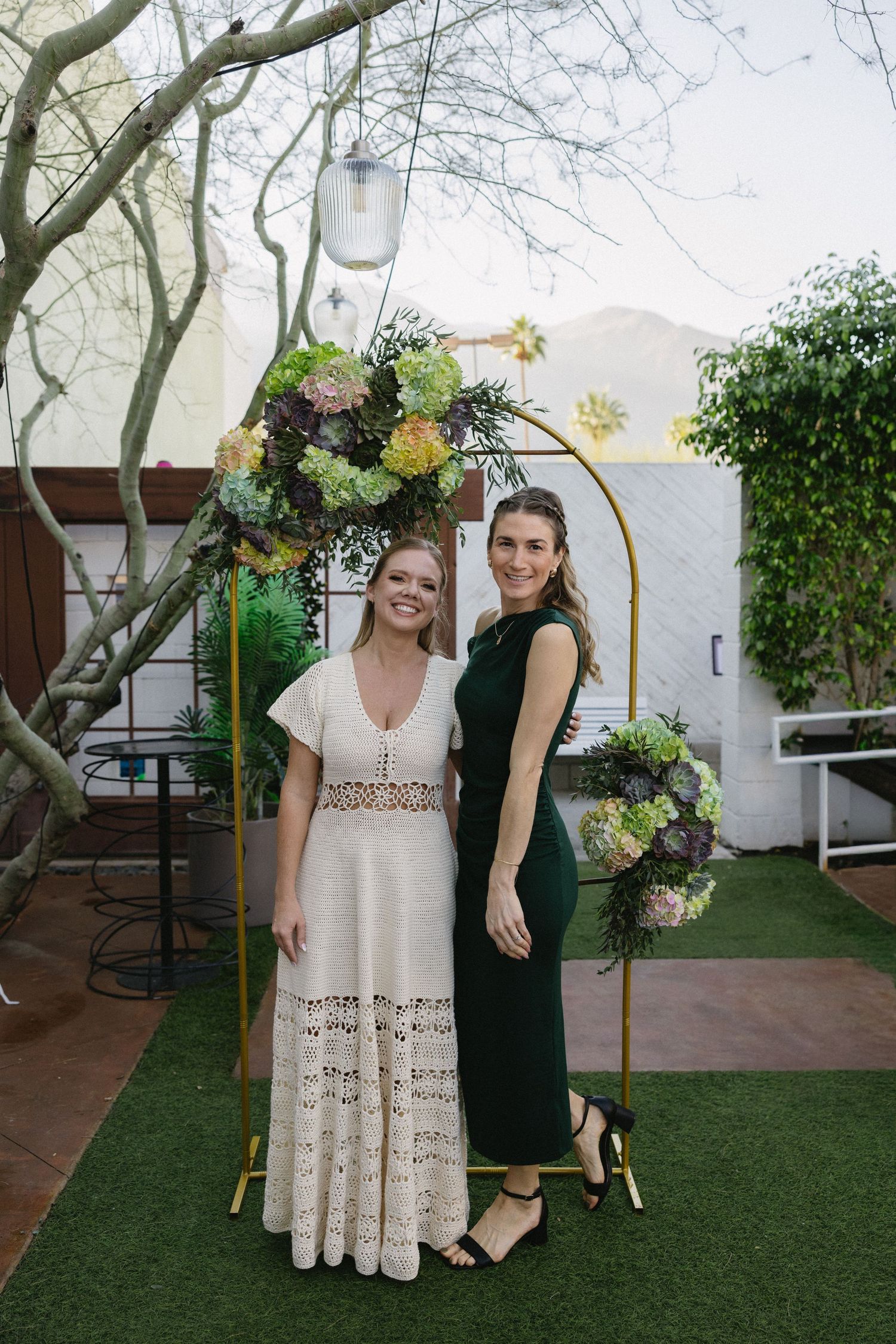 Two women posing under a gold arch with succulent floral arrangements at an outdoor wedding.