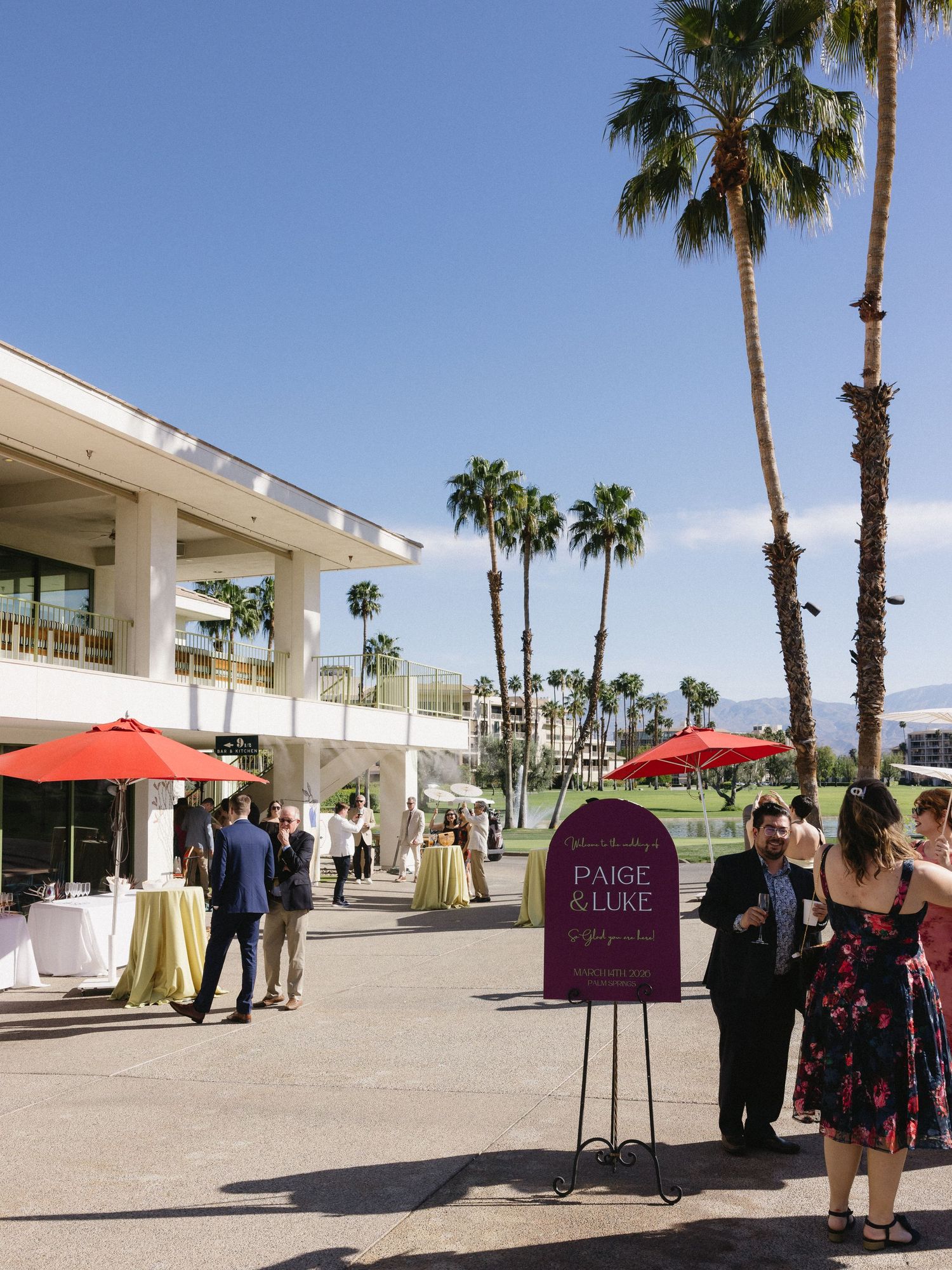 Outdoor wedding cocktail hour at a Palm Springs venue with palm trees, red umbrellas, and a Paige & Luke welcome sign.