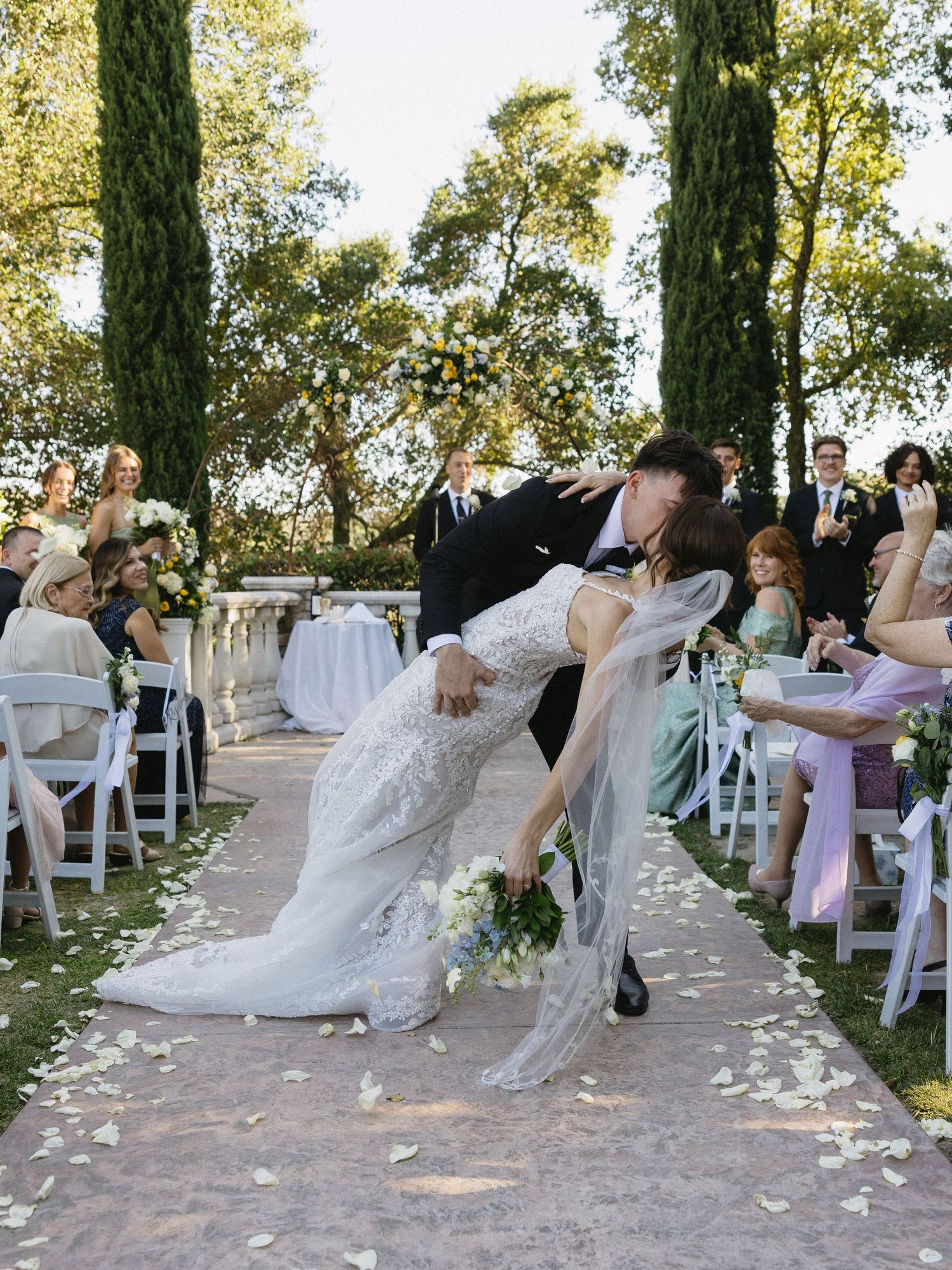 Groom dips bride for a kiss at outdoor wedding ceremony aisle lined with white chairs and rose petals.