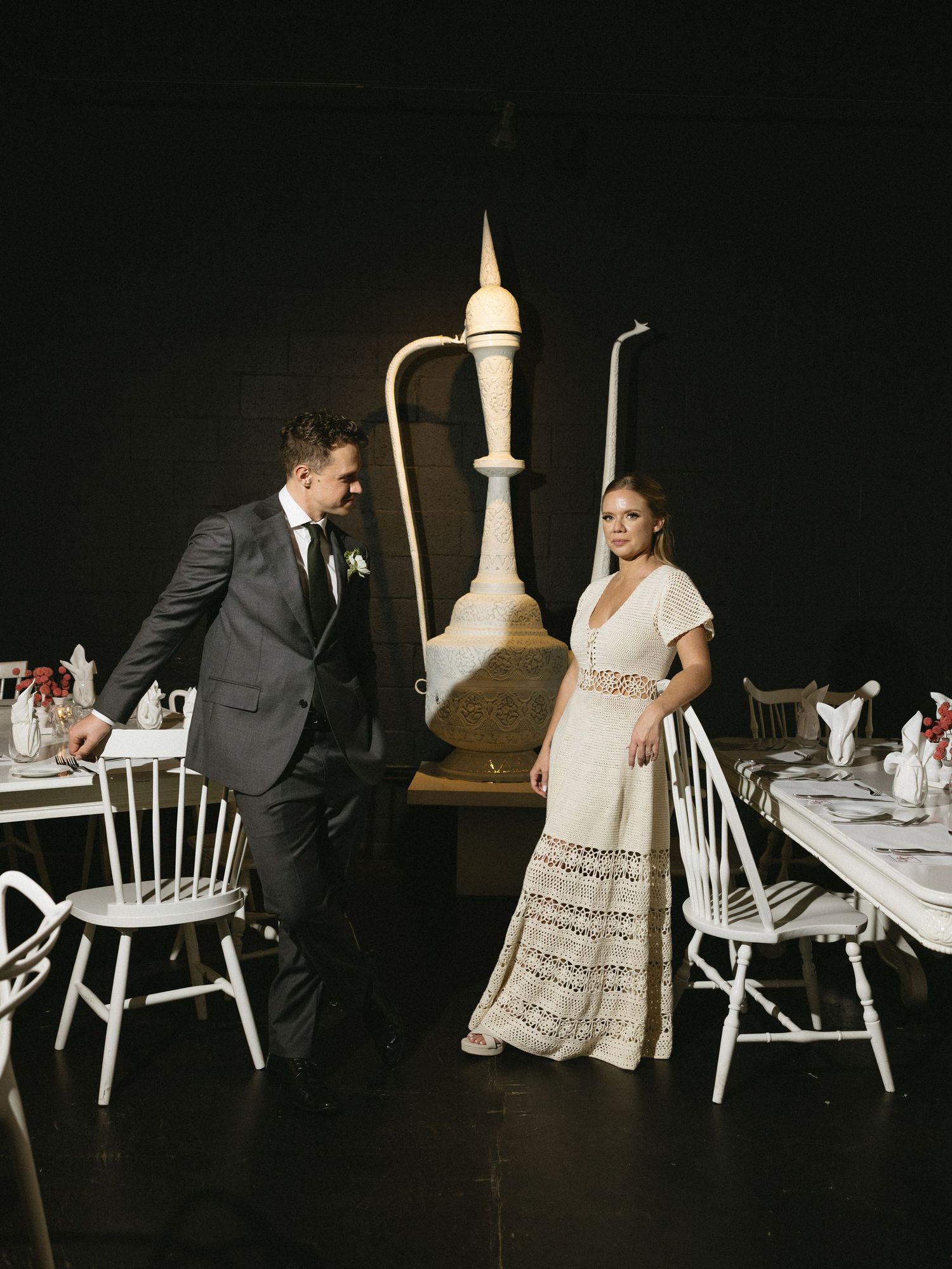 Bride in lace gown and groom in dark suit stand among white chairs at elegant black-walled wedding reception venue.