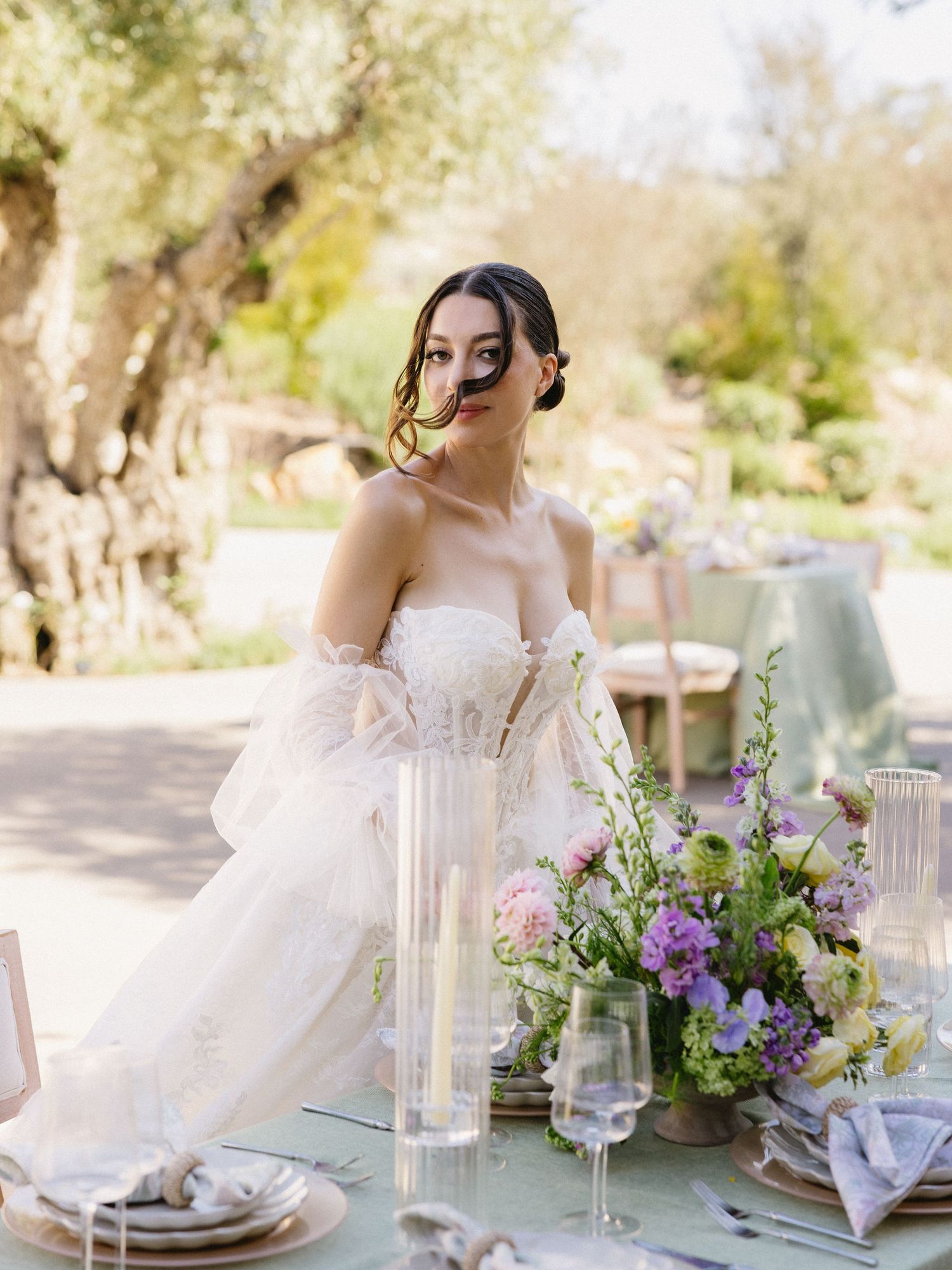 Bride in strapless white gown seated at elegant outdoor wedding reception table with pastel floral centerpiece.