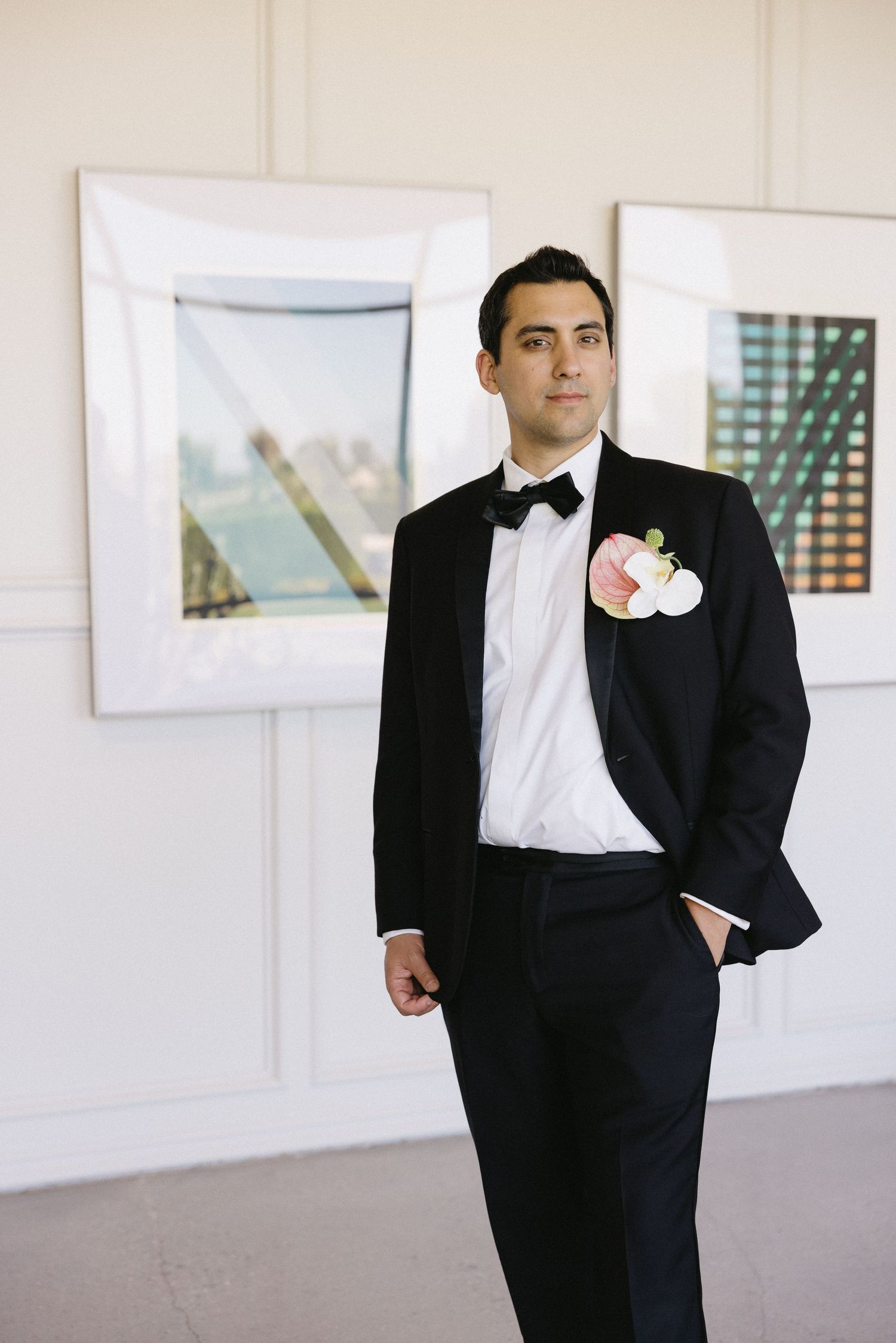 Groom in black tuxedo with bow tie and floral boutonniere standing in front of framed artwork.