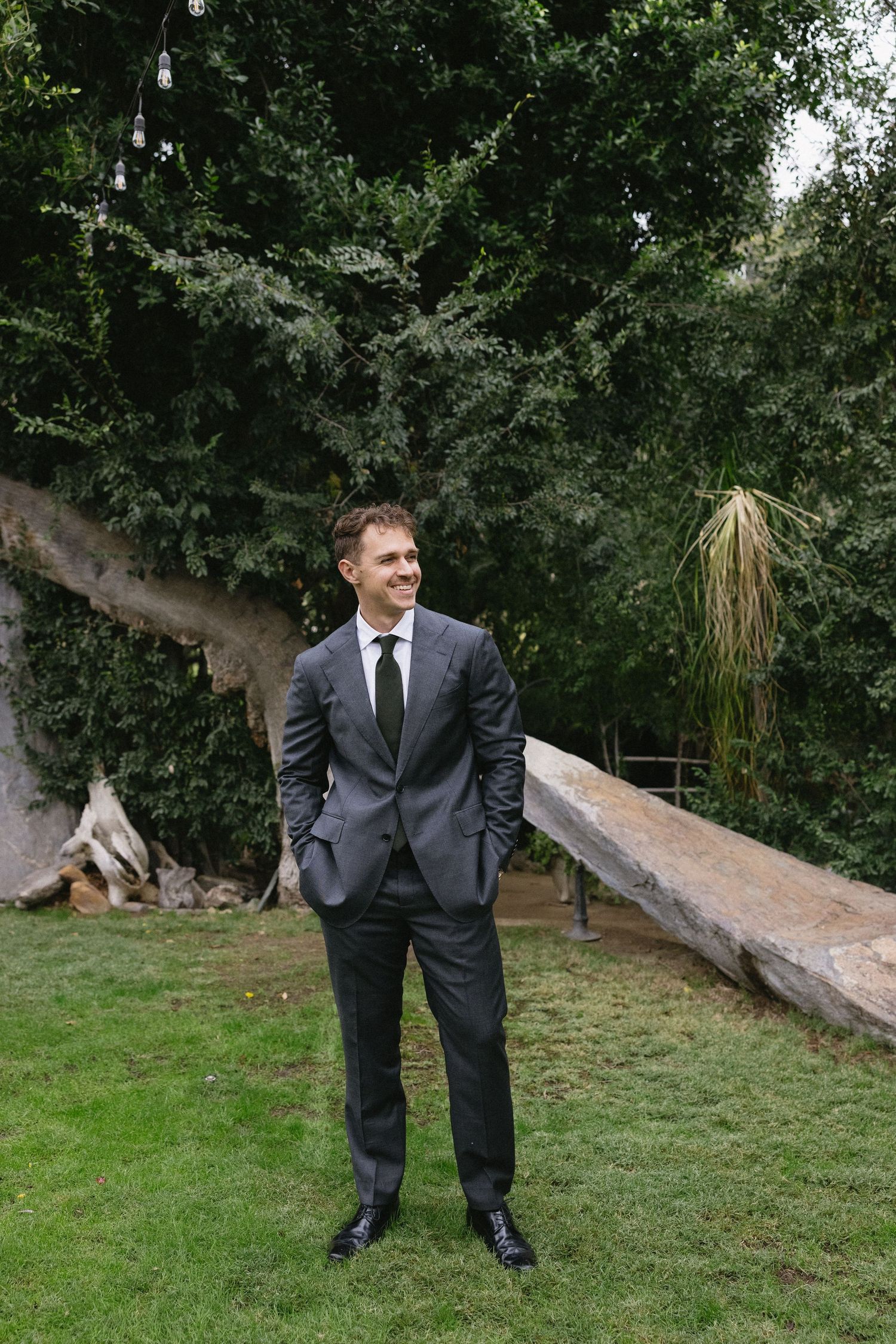 Groom in gray suit smiling outdoors before wedding ceremony.