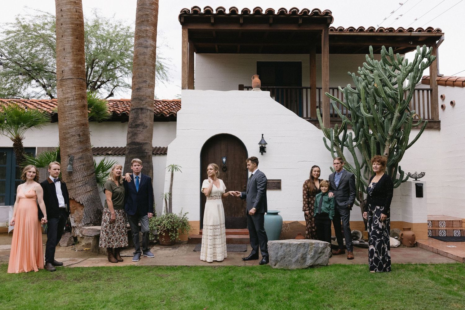 Wedding party poses outside a Spanish-style white stucco villa with cacti and palm trees in a desert setting.