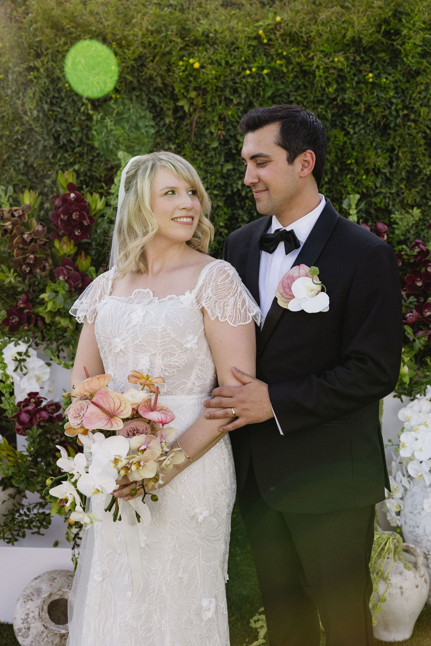 A smiling bride in lace dress holding floral bouquet stands with groom in black tuxedo outdoors at garden wedding.
