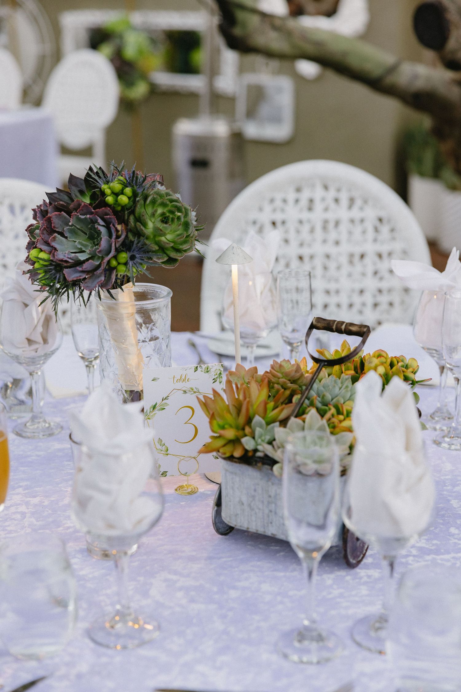 Elegant wedding table setting with succulent centerpieces, crystal glassware, white chairs, and candles on a lace tablecloth.