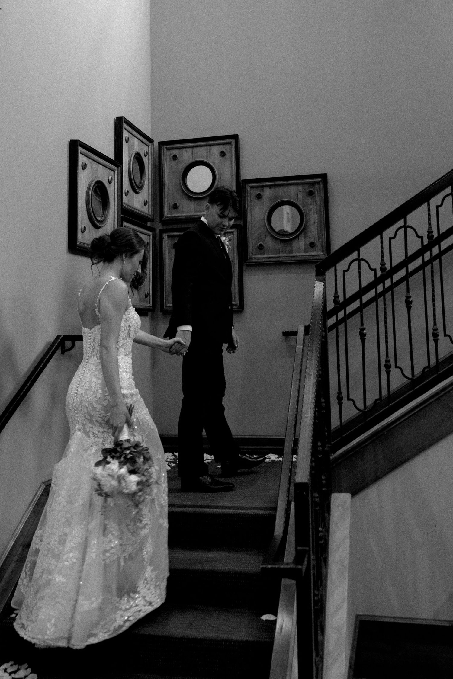 Black and white photo of a bride in lace gown holding bouquet, walking up stairs hand-in-hand with groom at wedding venue.