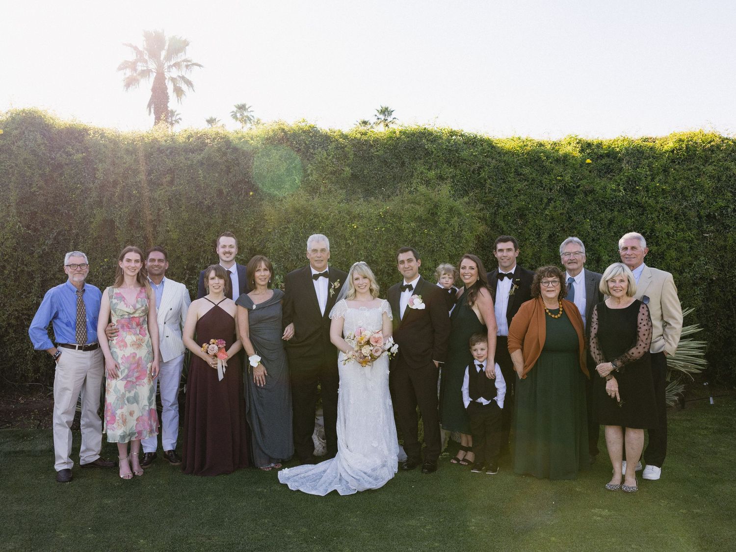Wedding party group photo outdoors on green lawn with bride and groom surrounded by family in formal attire.