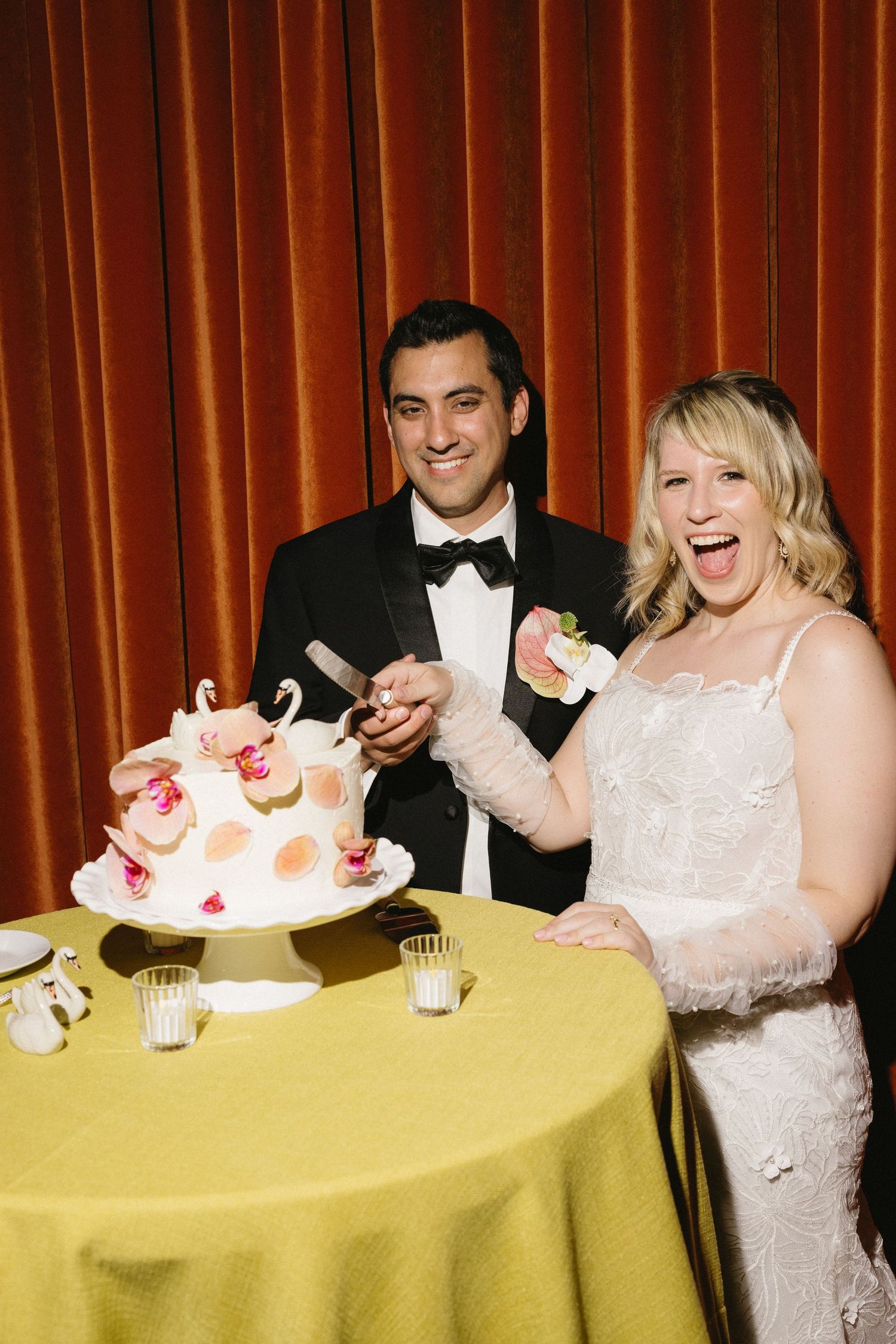 A joyful bride and groom cutting their wedding cake together at a reception, decorated with swan toppers on a yellow tablecloth.