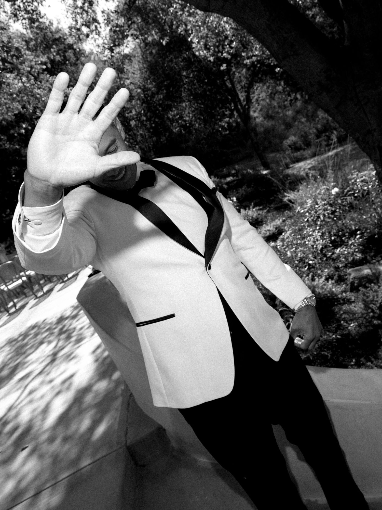 Man in white tuxedo jacket raises hand toward camera in black and white outdoor photo.