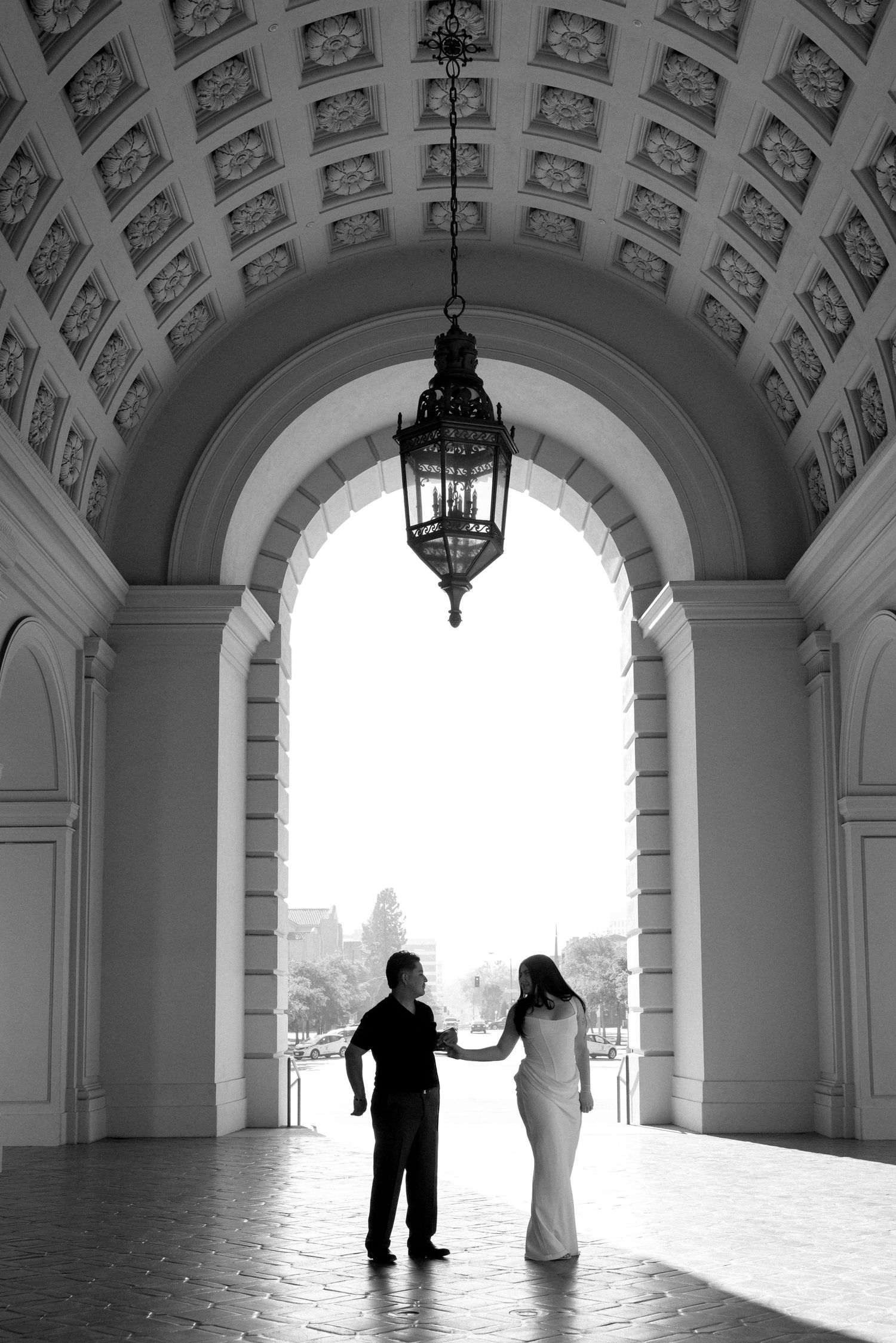 Black and white photo of a couple holding hands under an ornate arched entrance with a hanging lantern.