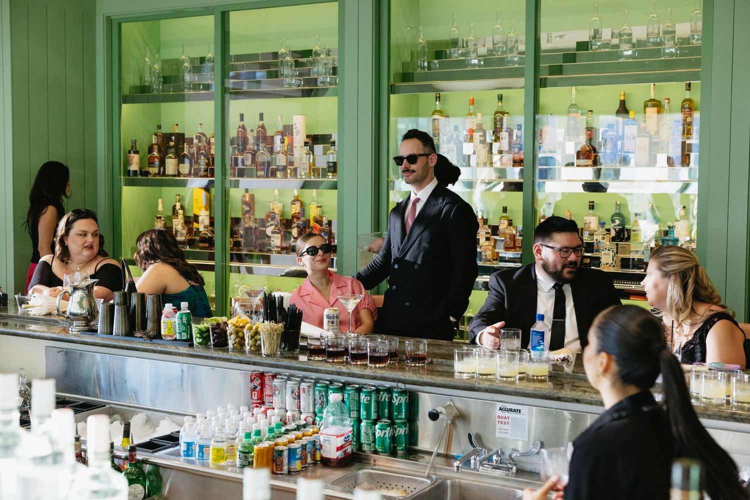 Bartender in sunglasses serves guests at a vibrant green bar stocked with colorful bottles and mixers.