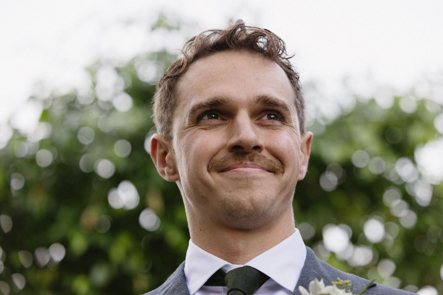 Smiling groom in suit with boutonniere looks up joyfully outdoors at wedding ceremony.
