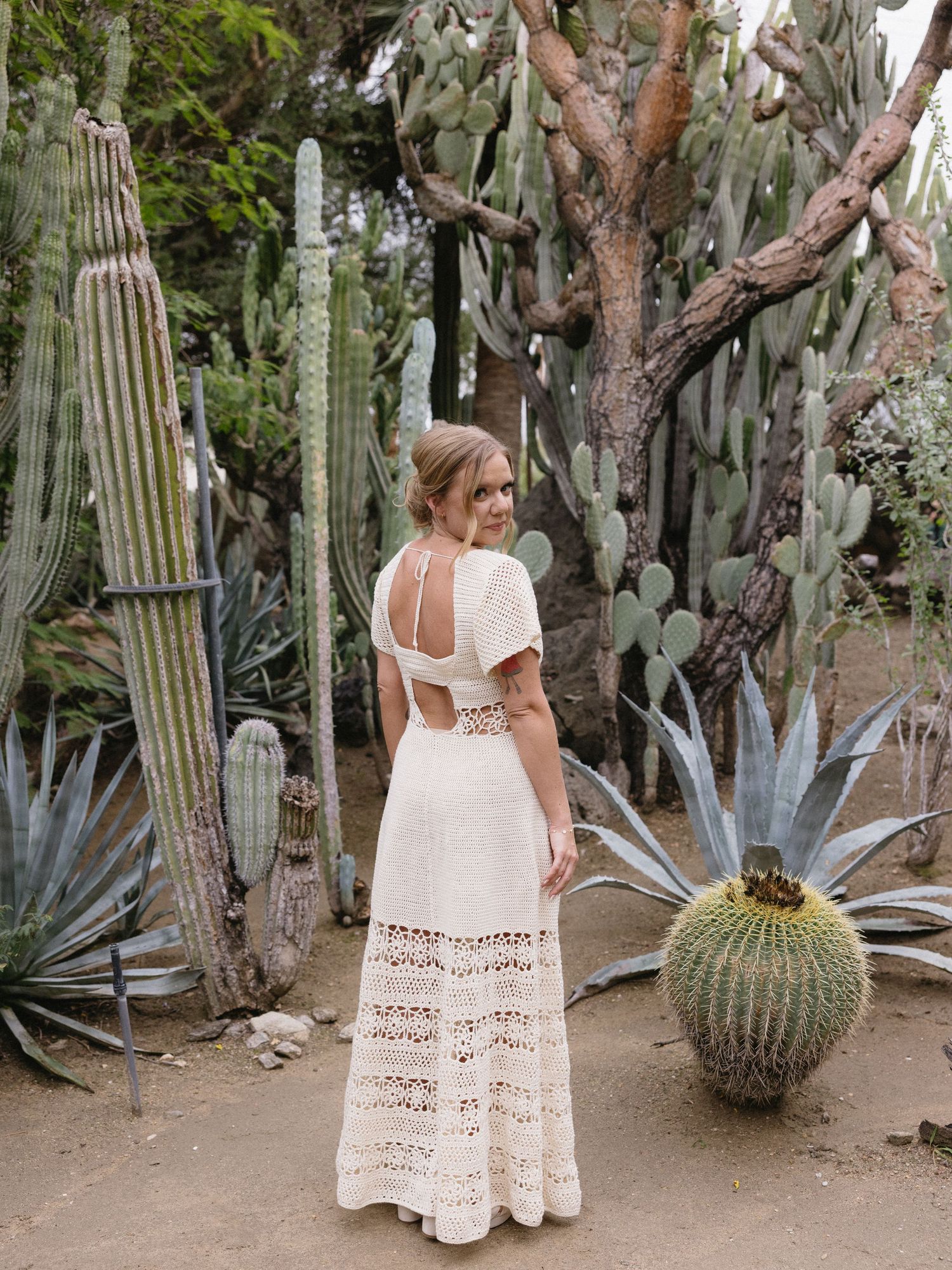 Woman in white crochet backless maxi dress posing among tall cacti and succulents in a desert garden.