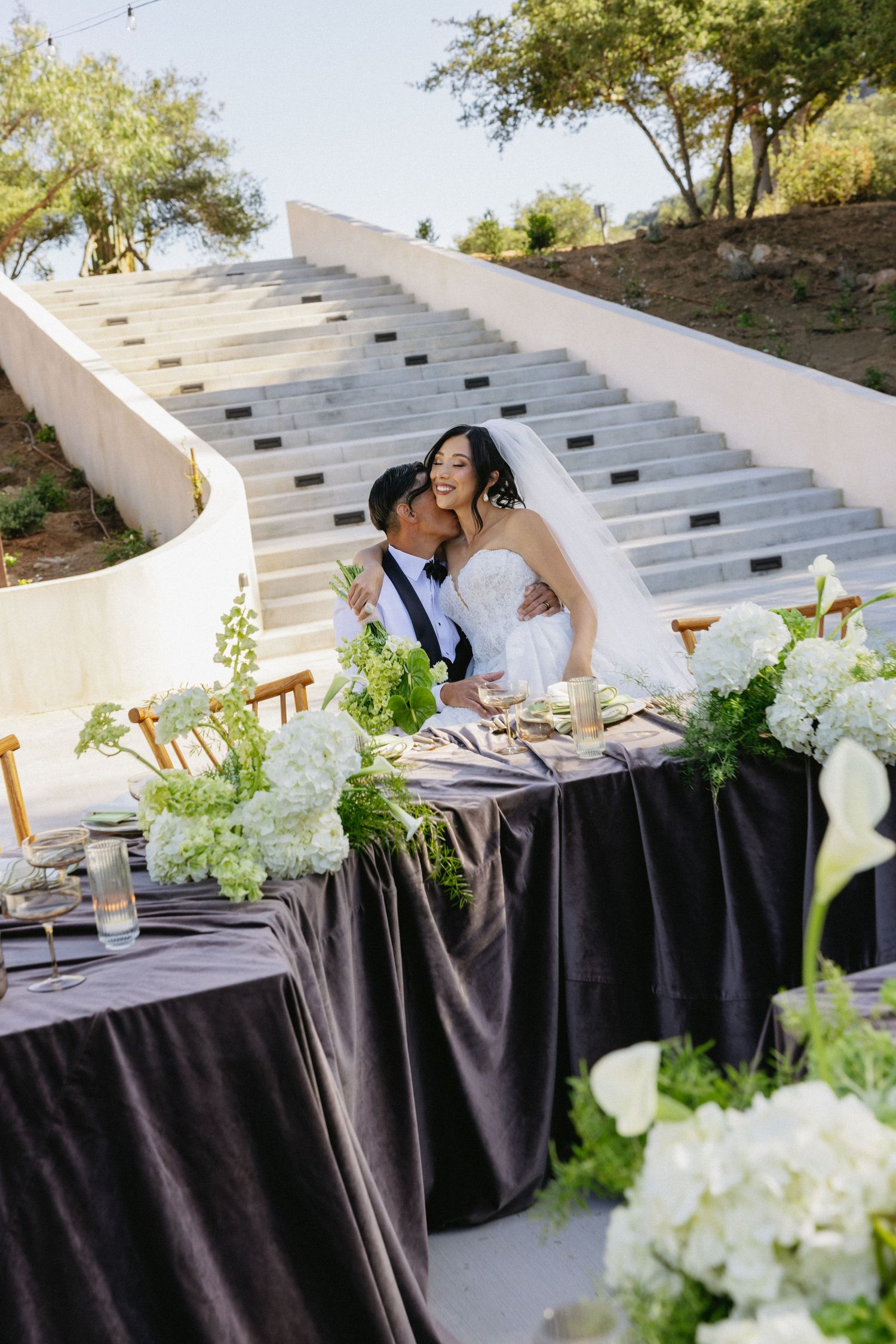 Bride and groom share a romantic kiss at outdoor wedding sweetheart table adorned with white hydrangeas and greenery.