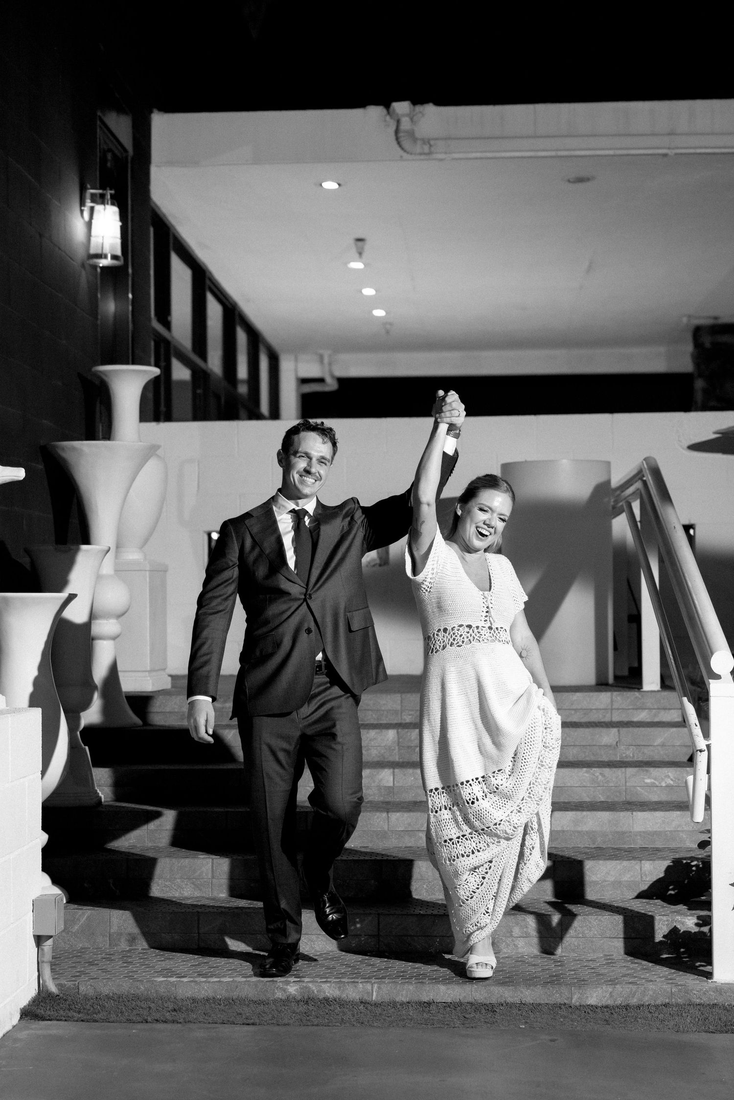 Black and white photo of a joyful bride and groom descending stairs hand in hand at their wedding reception.