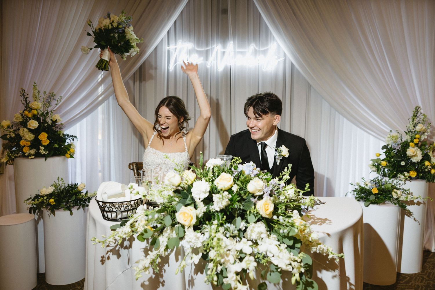 Joyful bride and groom celebrate at wedding reception sweetheart table with white and yellow floral arrangements.