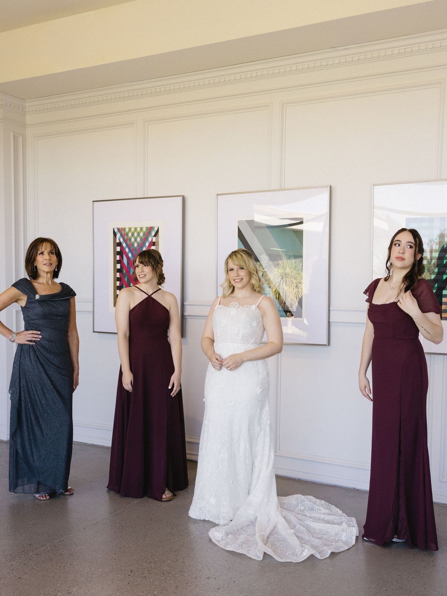Bride in white lace gown poses with bridesmaids in burgundy and navy dresses in elegant gallery venue.