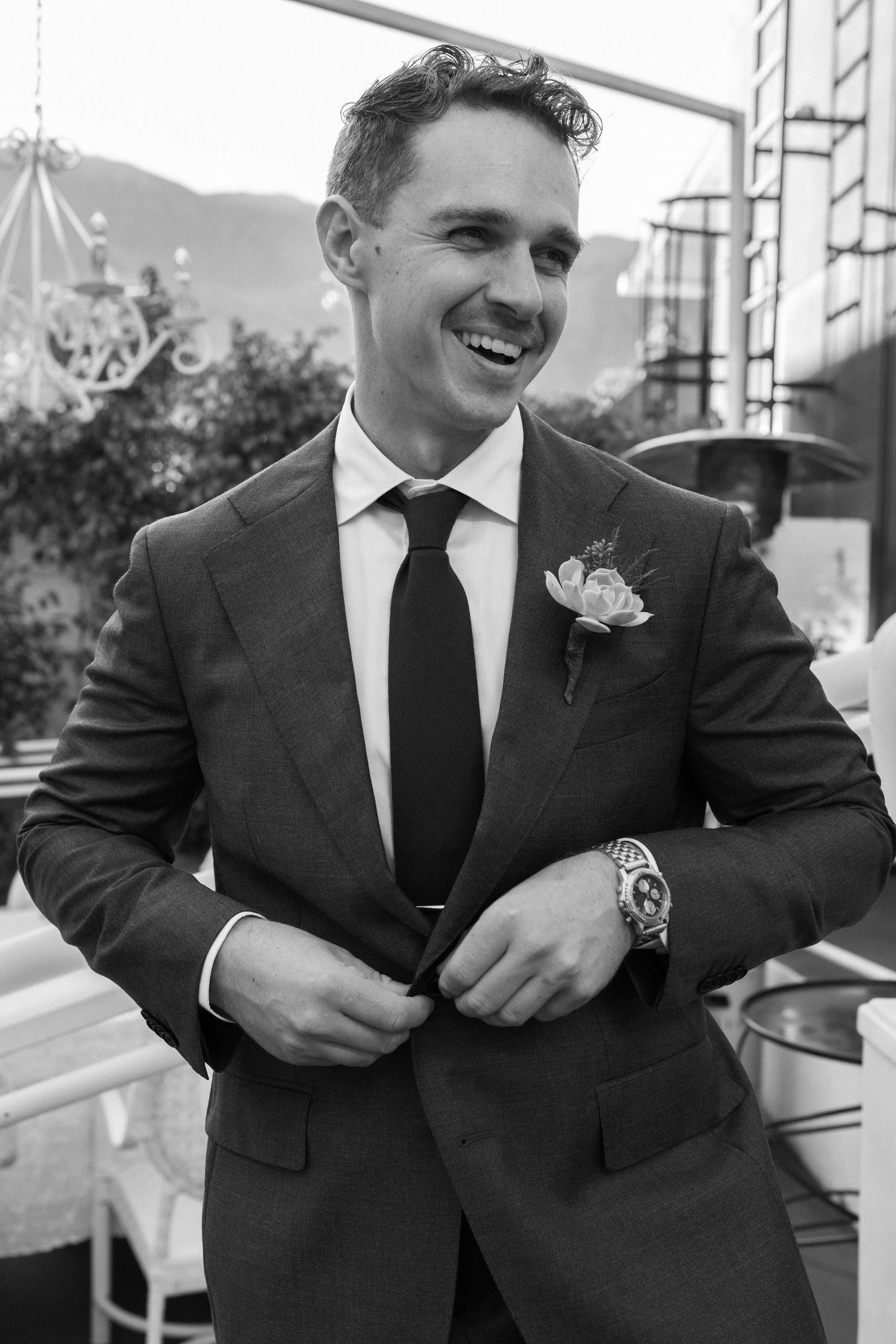 Smiling groom in a tailored suit with boutonniere adjusts his jacket button outdoors in a black and white photo.