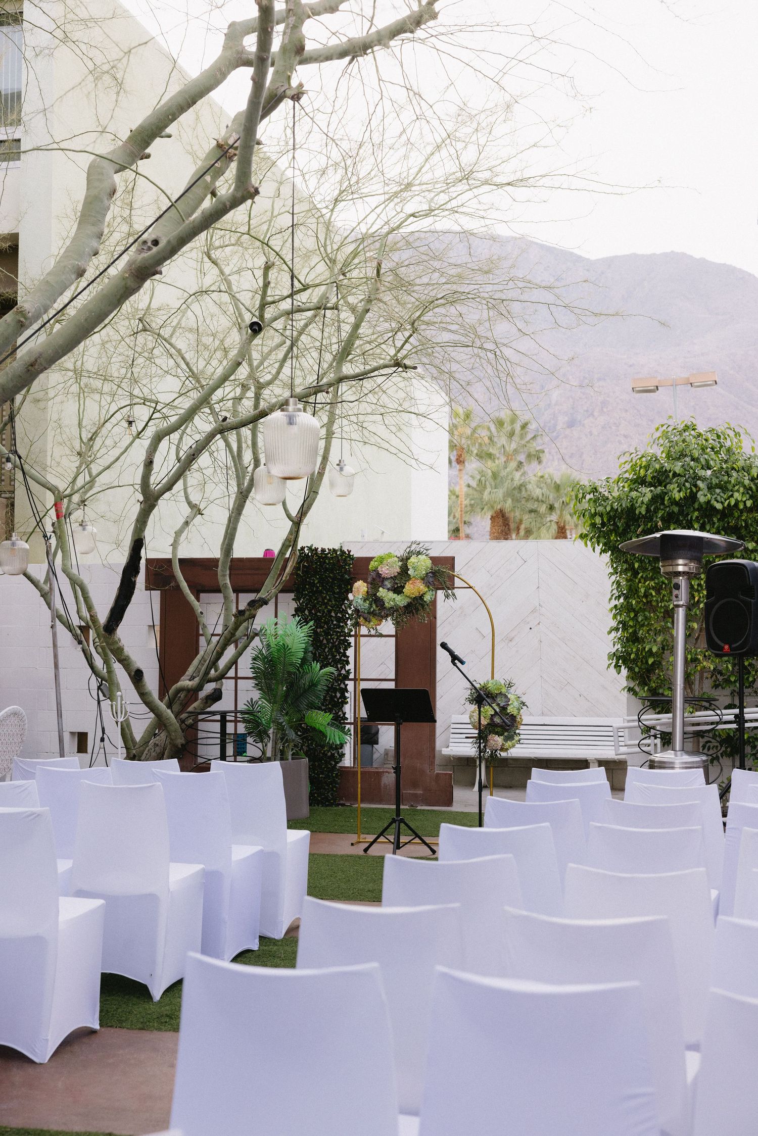 Outdoor wedding ceremony setup with white chair covers, floral arch, bare tree, and mountain views.