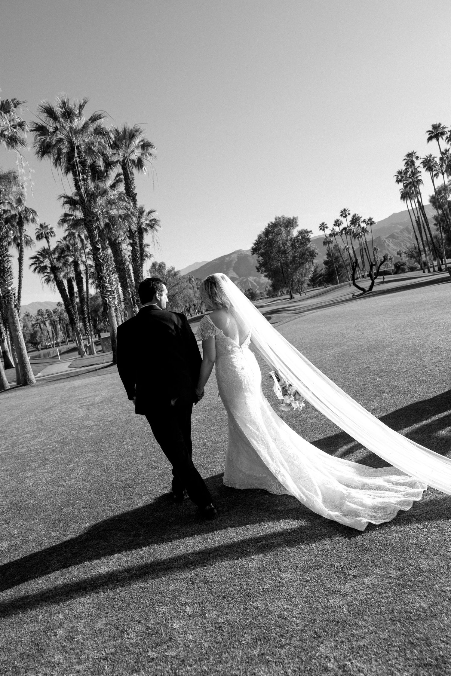 Black and white photo of bride and groom walking hand in hand on a golf course with palm trees and mountains.